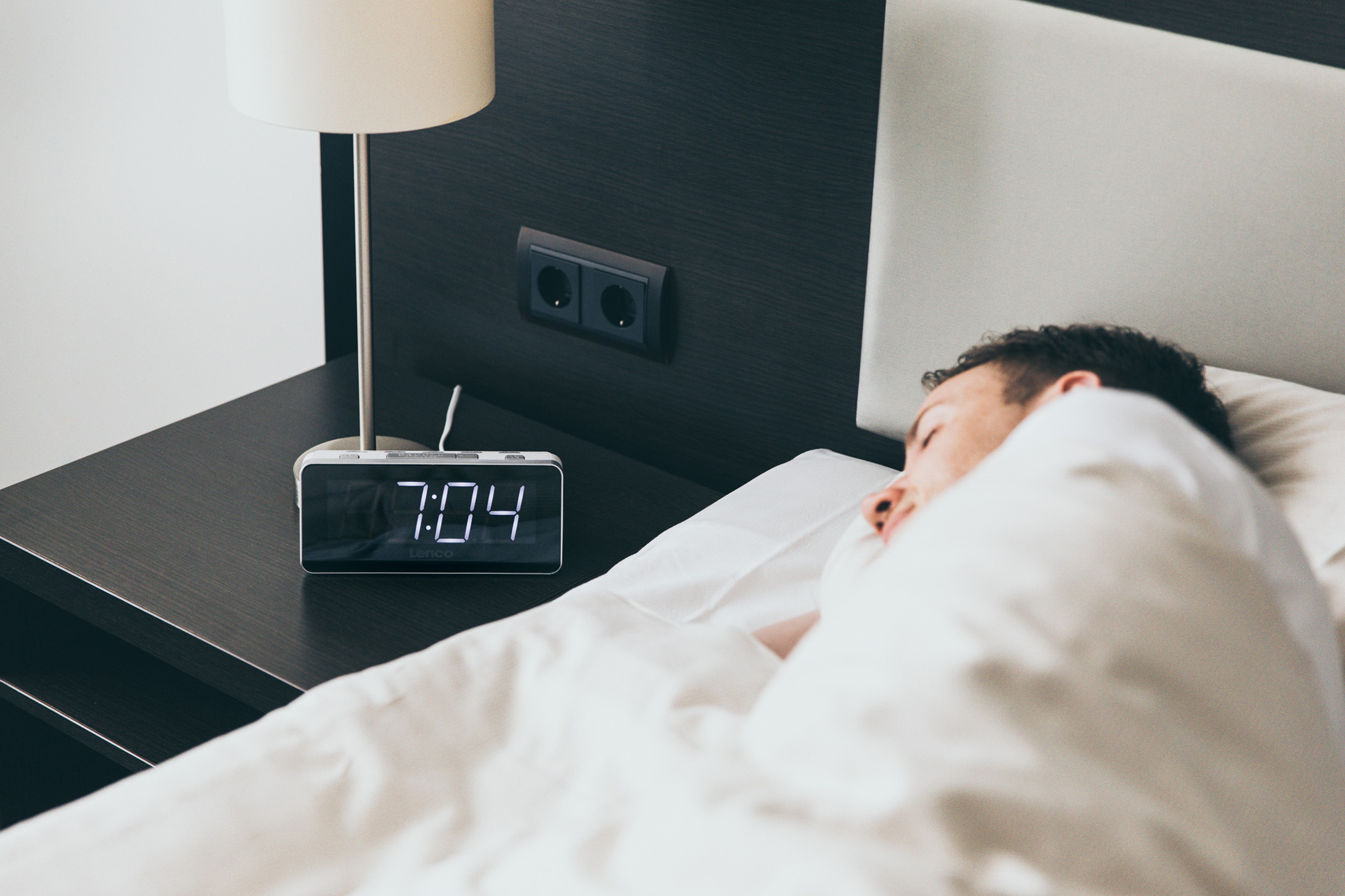 Osoba śpiąca w łóżku, z budzikiem pokazującym 7:04 na stoliku nocnym.