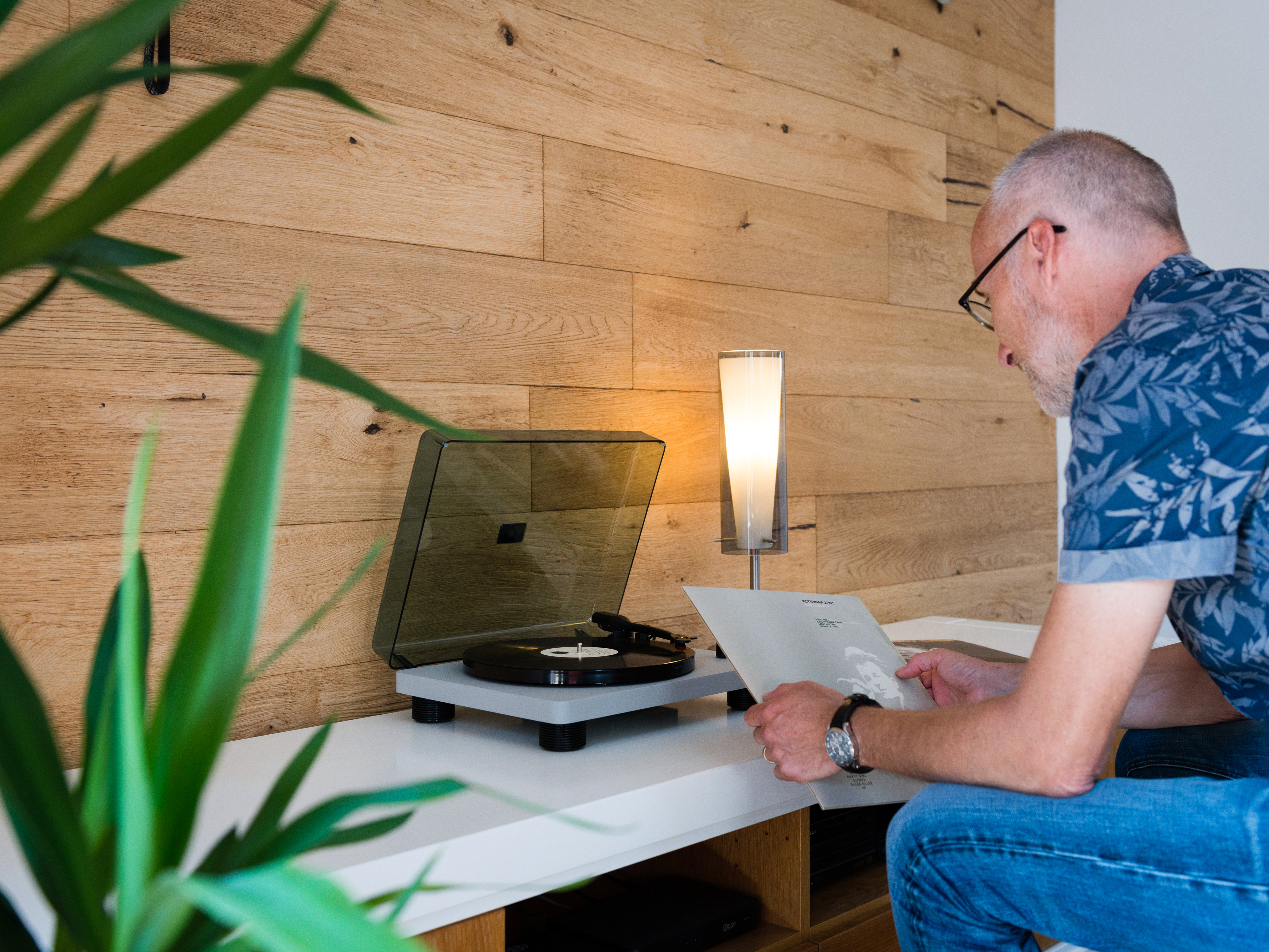 Mężczyzna w okularach i koszuli ogląda płytę obok gramofonu i lampy.