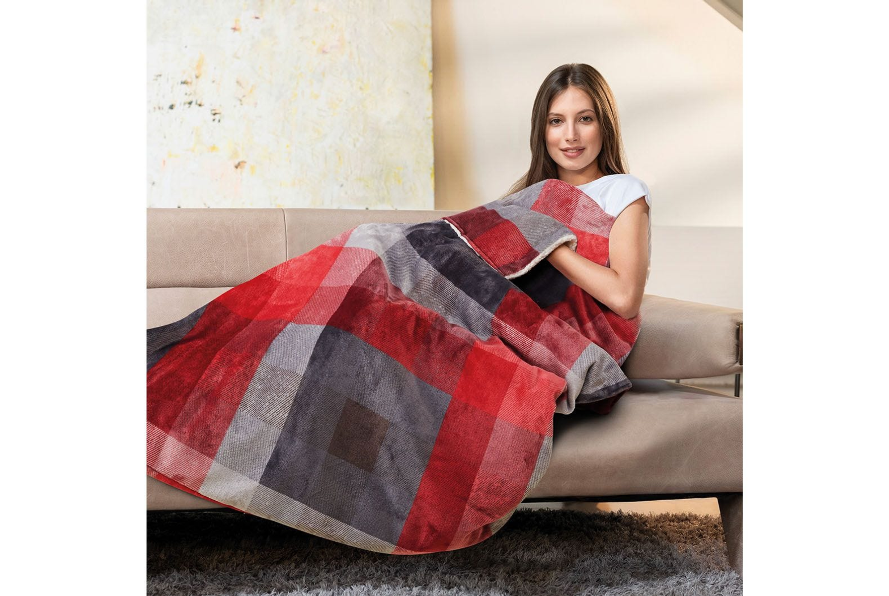 Donna su un divano coperta da una coperta a quadri rossa e grigia. Sorride e guarda la fotocamera.