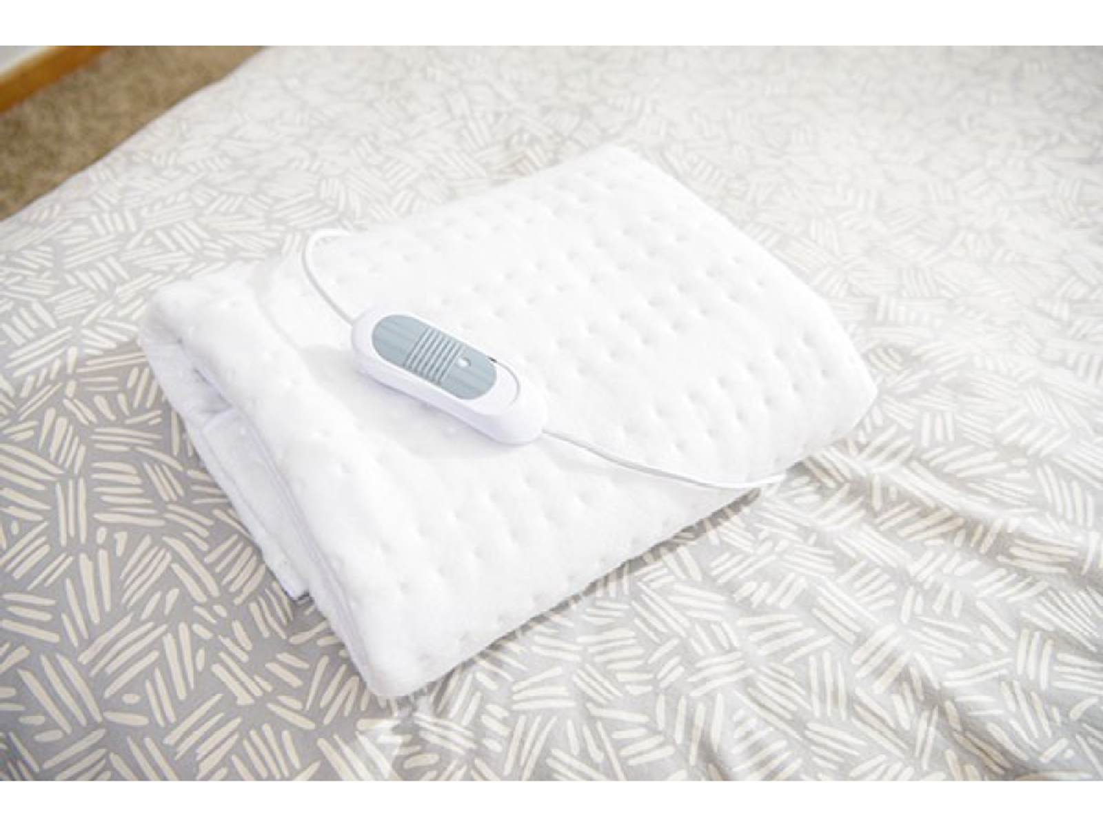 Manta térmica eléctrica blanca doblada en una cama con sábanas grises estampadas.