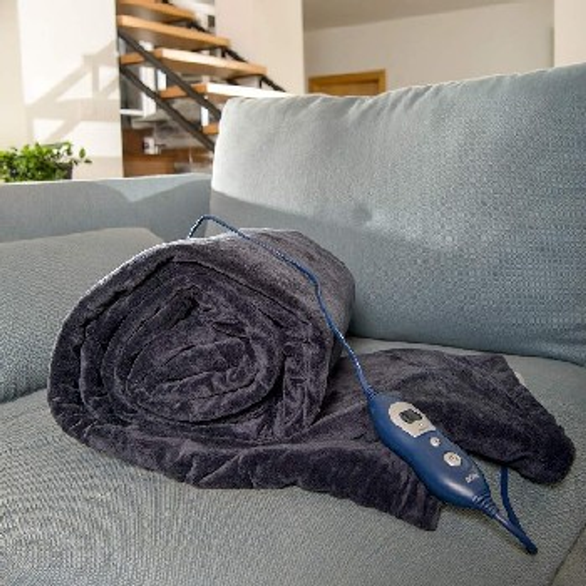 Zwinięty granatowy koc elektryczny z jednostką sterującą. Leży na niebieskiej kanapie. Jasne tło.