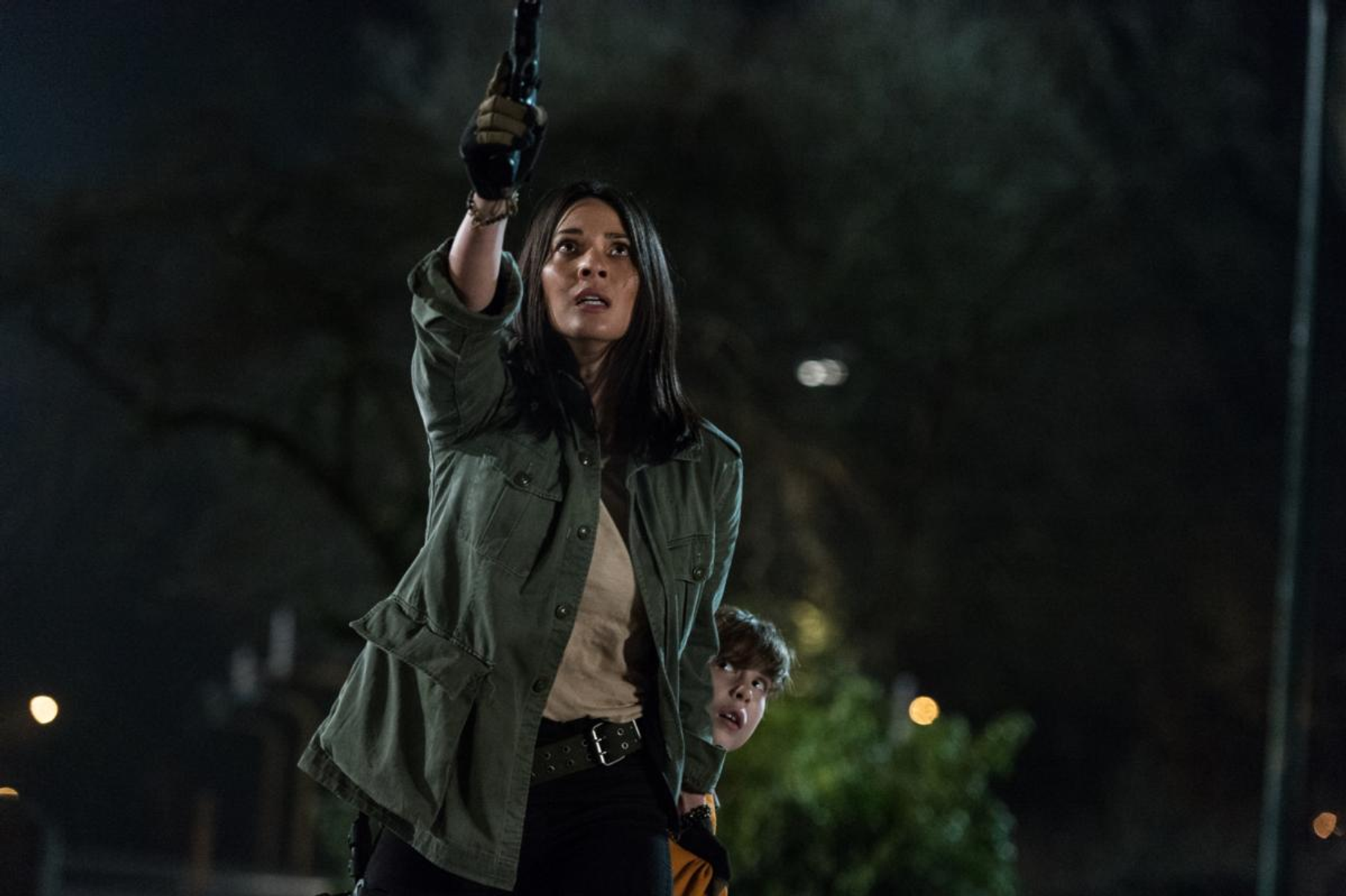 Imagen: Mujer sosteniendo un arma, un niño detrás de ella. Escena nocturna oscura.