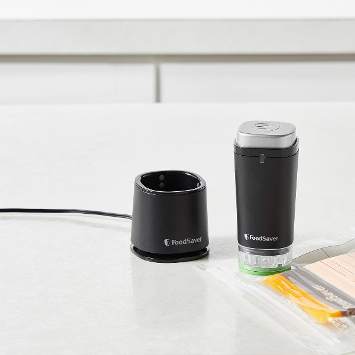 Sellador al vacío FoodSaver negro y accesorios sobre una superficie blanca. Incluye una base de carga.