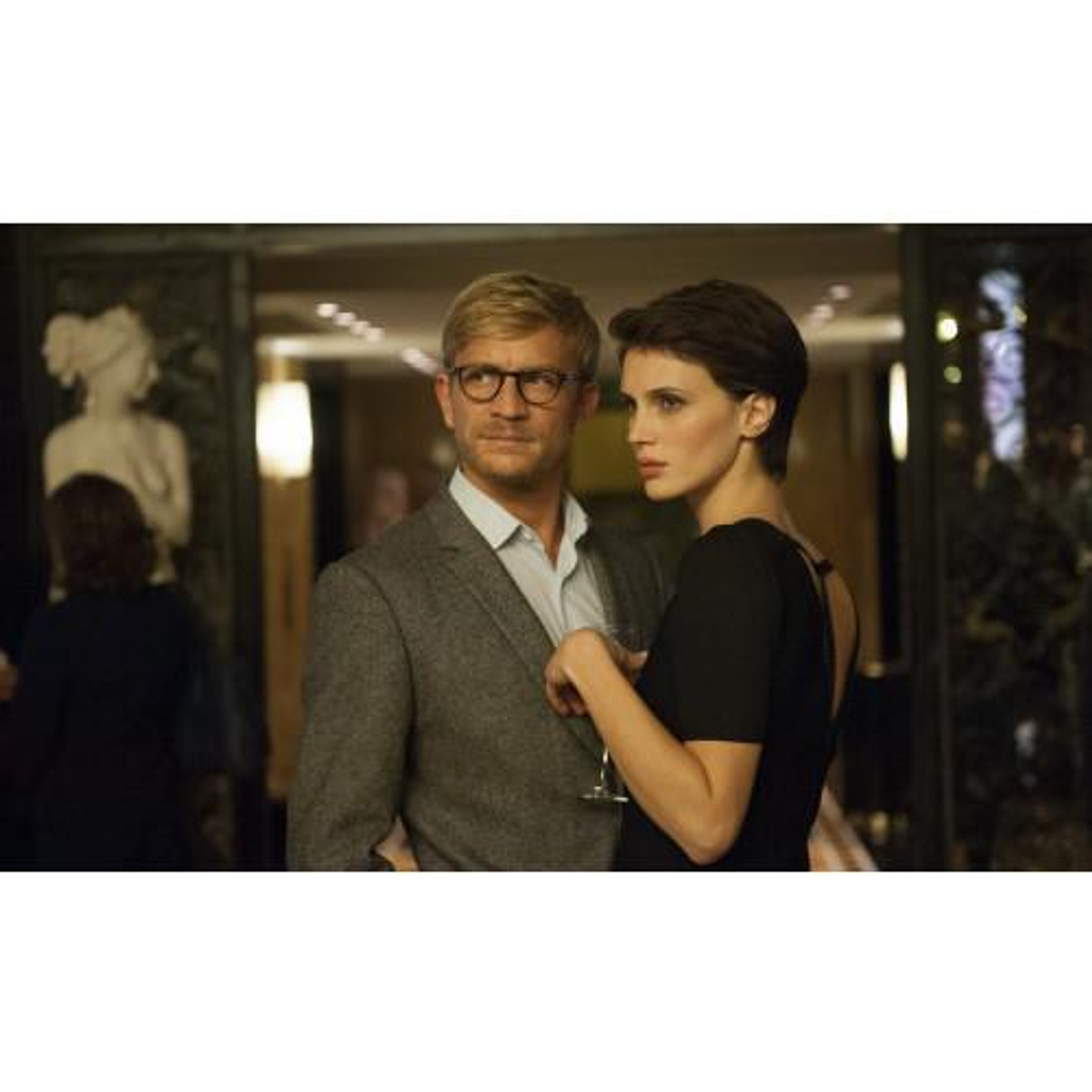 Escena de película: Hombre y mujer en un interior. Hombre con gafas y mujer con vestido negro.