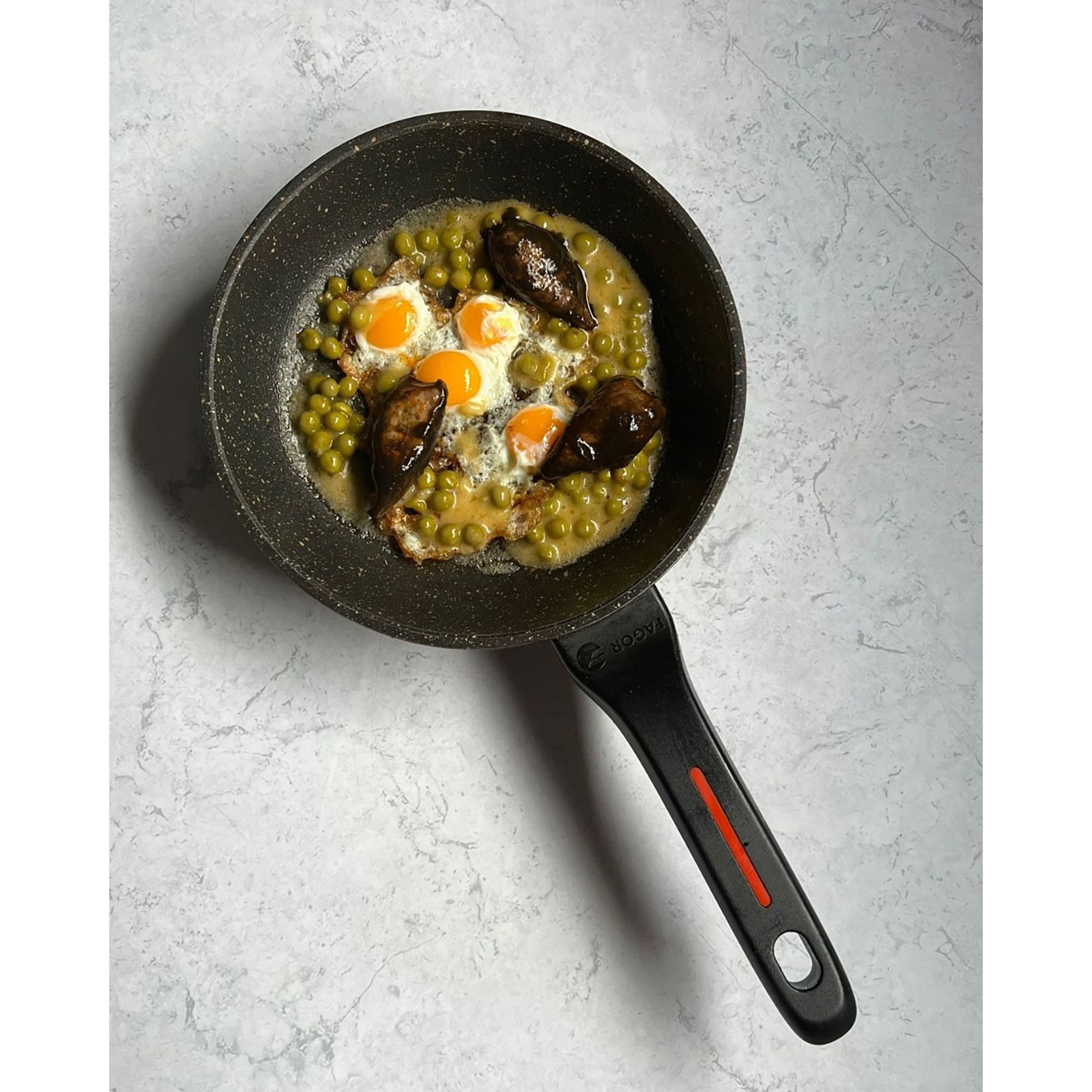 Sartén con huevos fritos, guisantes y carne. La sartén tiene un mango negro y está sobre mármol.