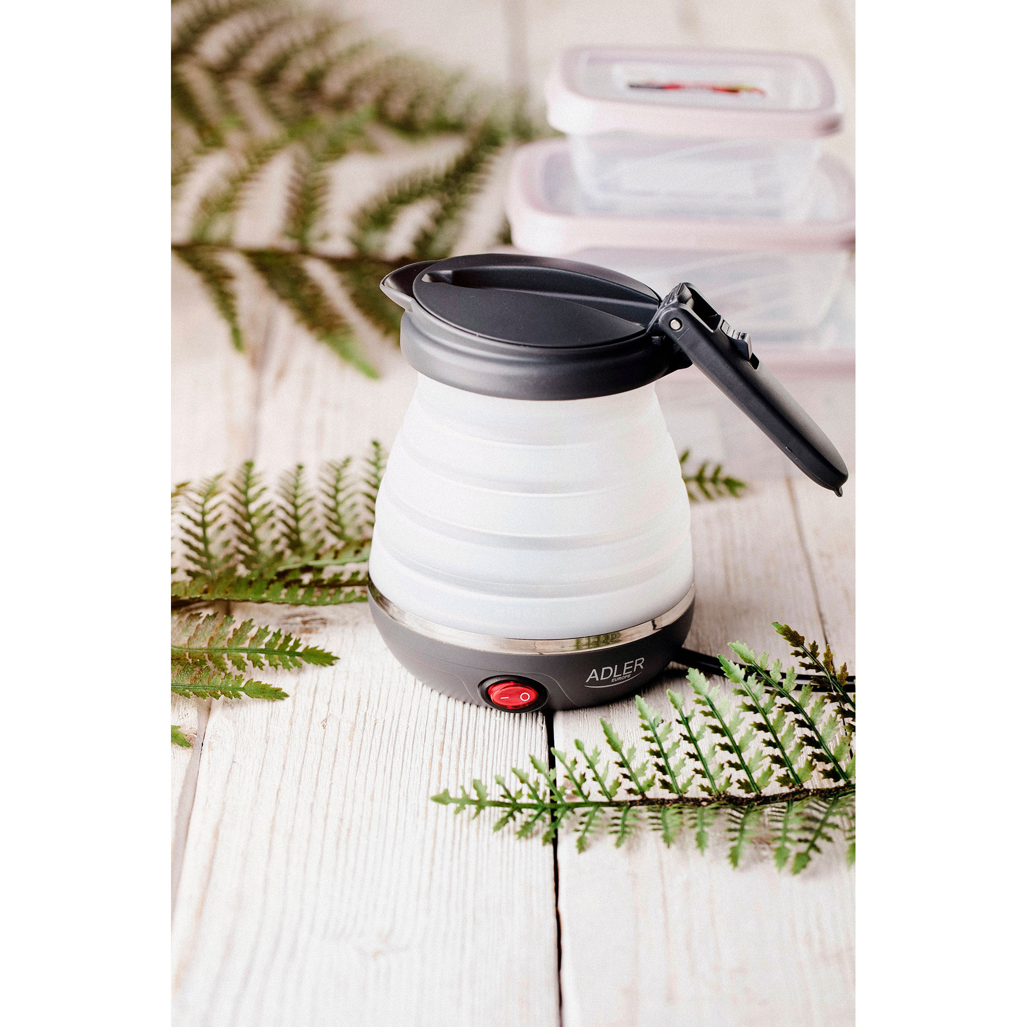 Składany czajnik z czerwonym przyciskiem zasilania, z liśćmi paproci w tle.