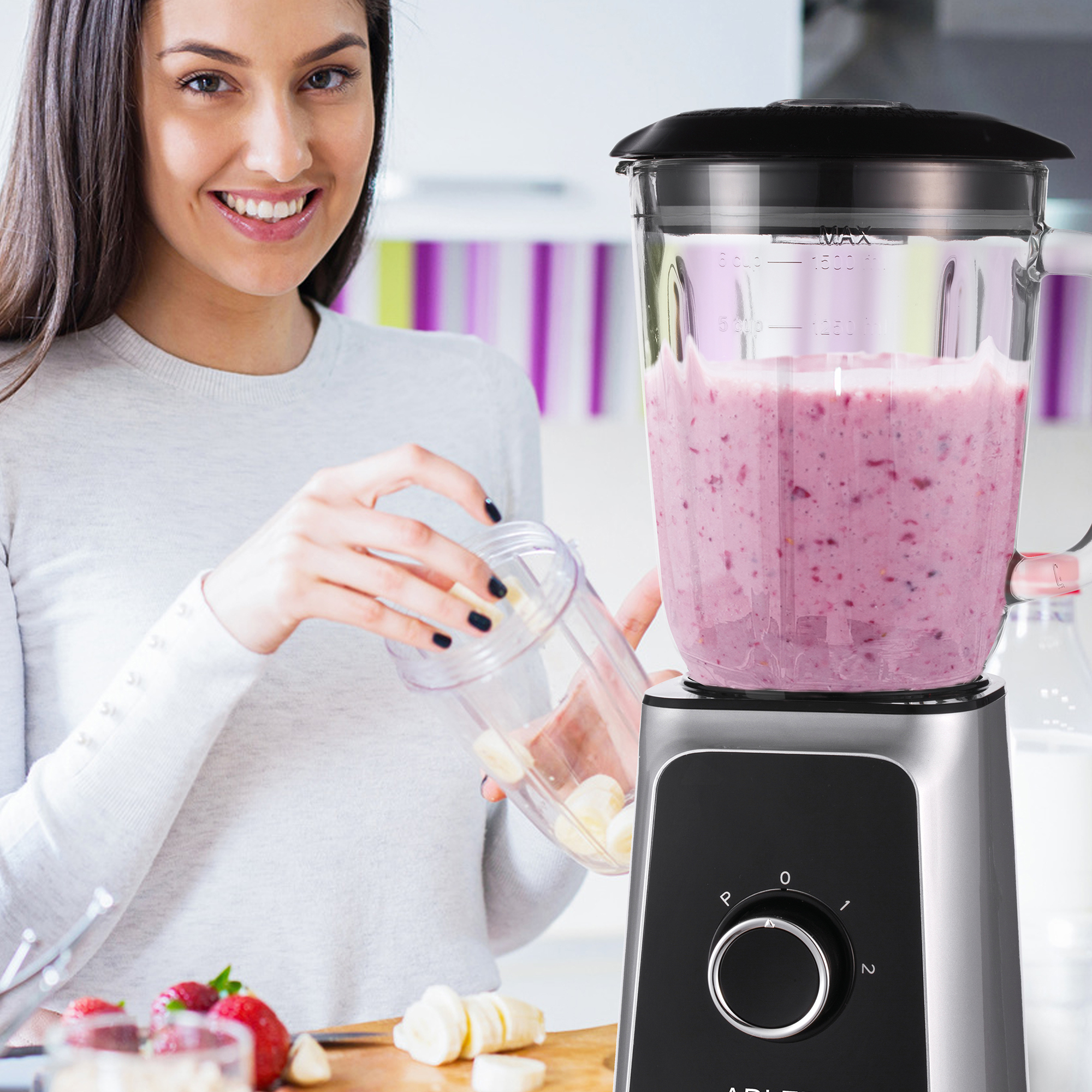 Kobieta z blenderem. Uśmiecha się i trzyma pojemnik. Blender ma różowe smoothie i banany.