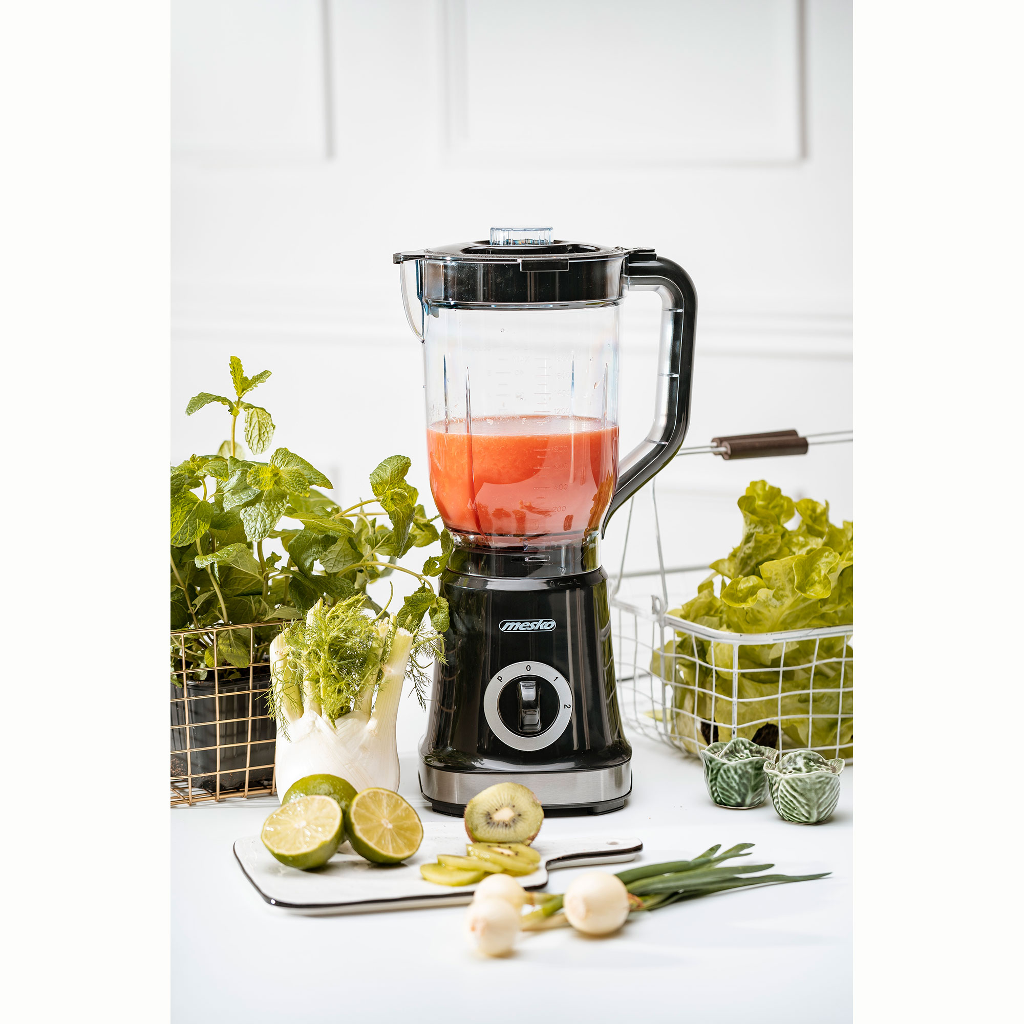 Blender z czerwoną cieczą w środku, otoczony składnikami: limonkami, kiwi, sałatą, ziołami i cebulą na białej powierzchni.