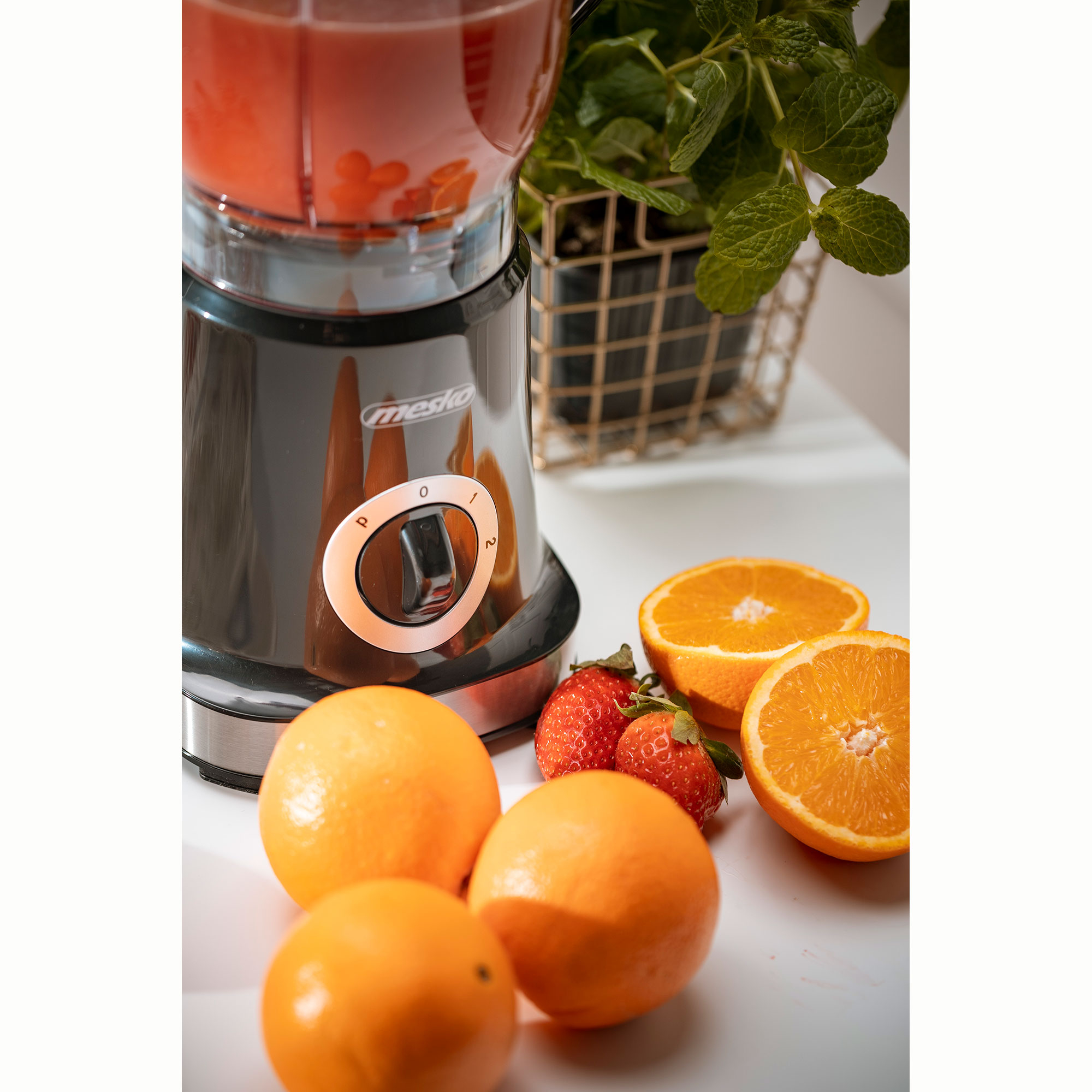 Blender z plasterkami pomarańczy, pomarańczami i truskawkami na białej powierzchni.