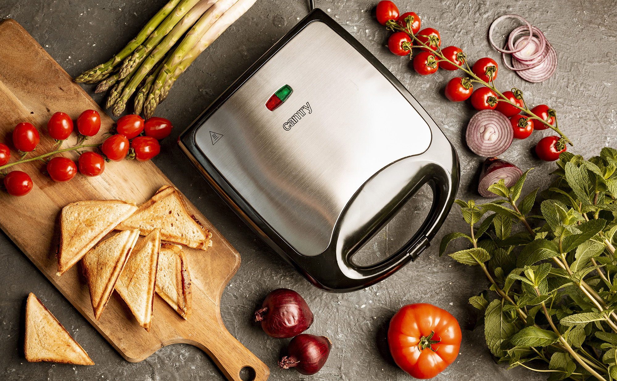 Sandwich maker with ingredients: tomatoes, asparagus, onions, mint, and bread. Gray background.