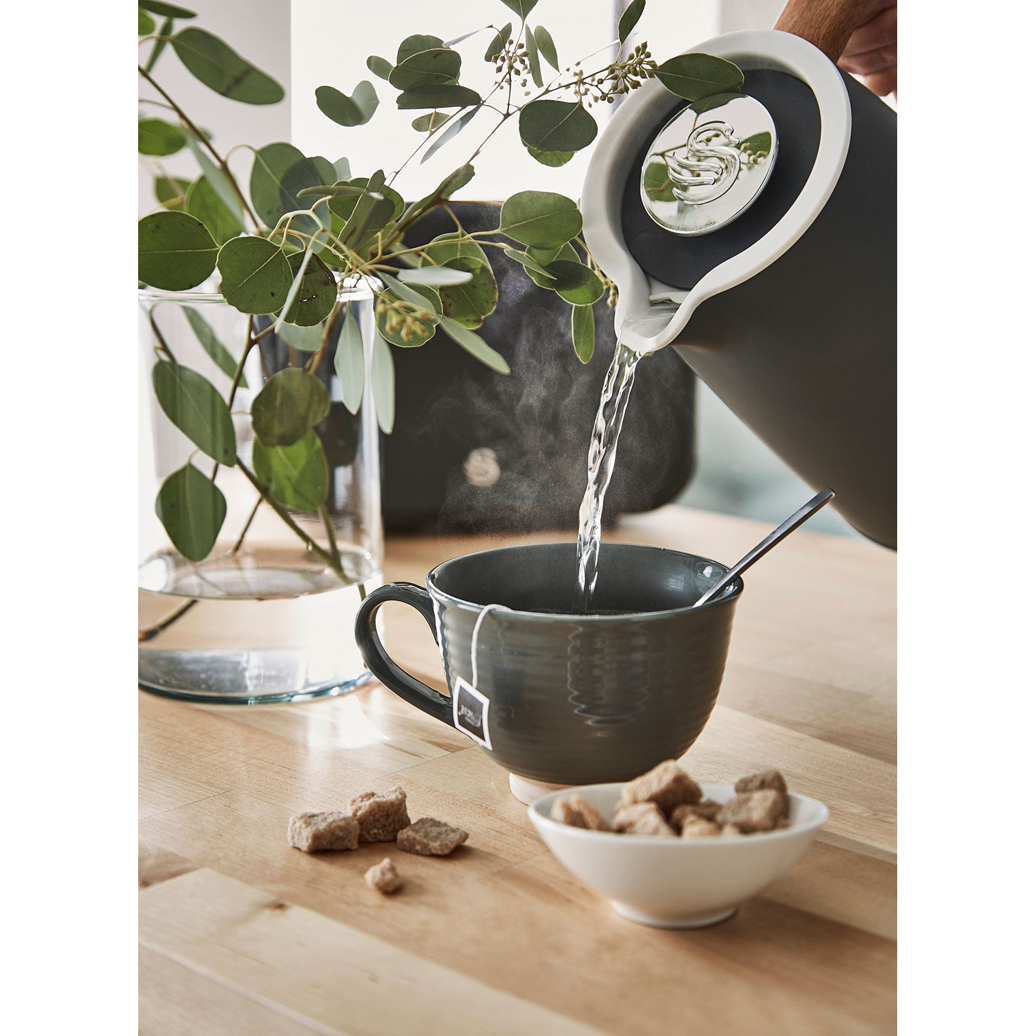 Eine Person gießt Wasser aus einem dunklen Wasserkocher in eine Tasse mit Teebeutel. Zucker und Blätter liegen auf dem Holztisch.