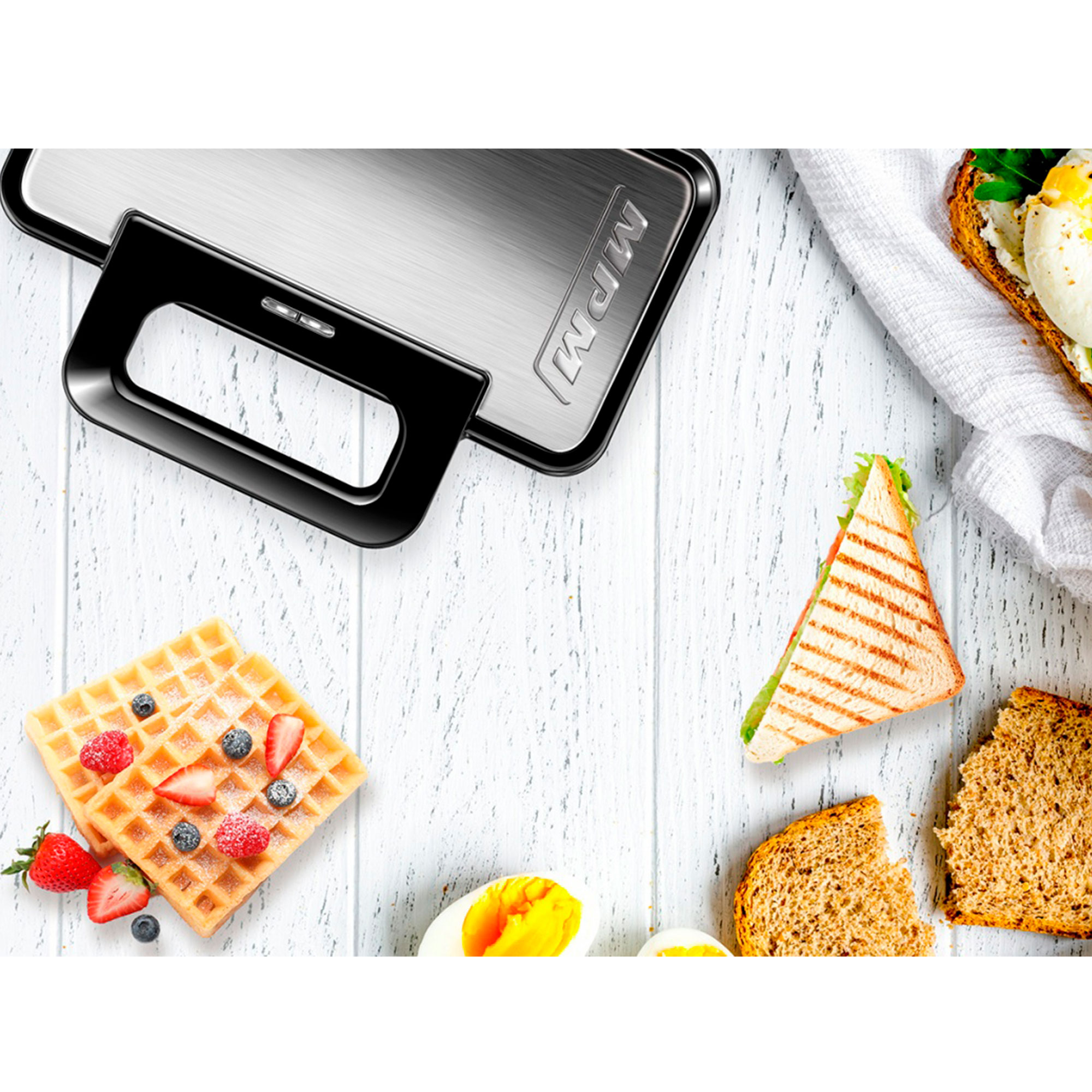 Gofrownica z uchwytem, gofry z jagodami, kanapka i kromki chleba na białym drewnianym stole.