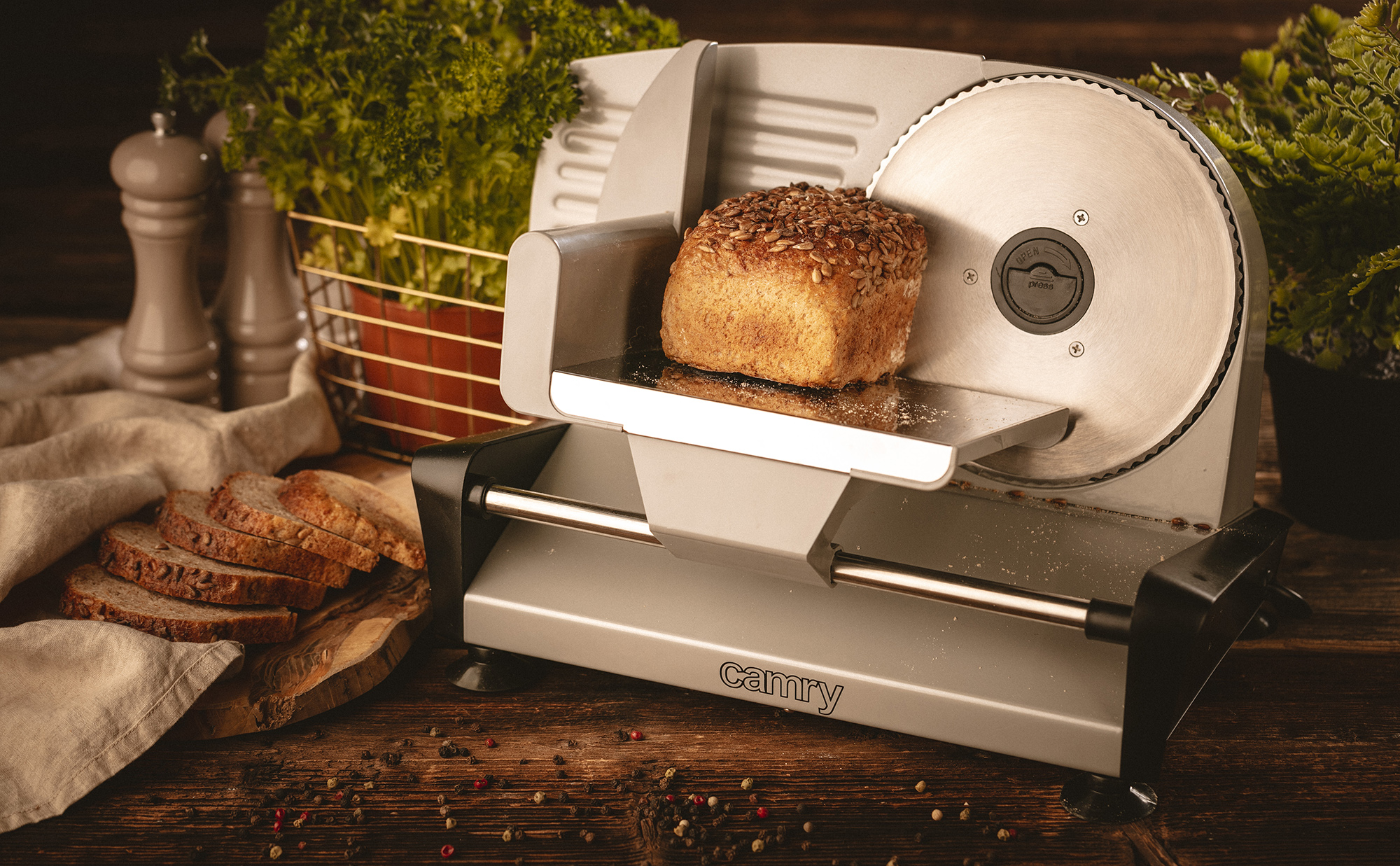 Krajalnica do chleba z bochenkiem na ostrzu, kromkami i zielenią w tle.