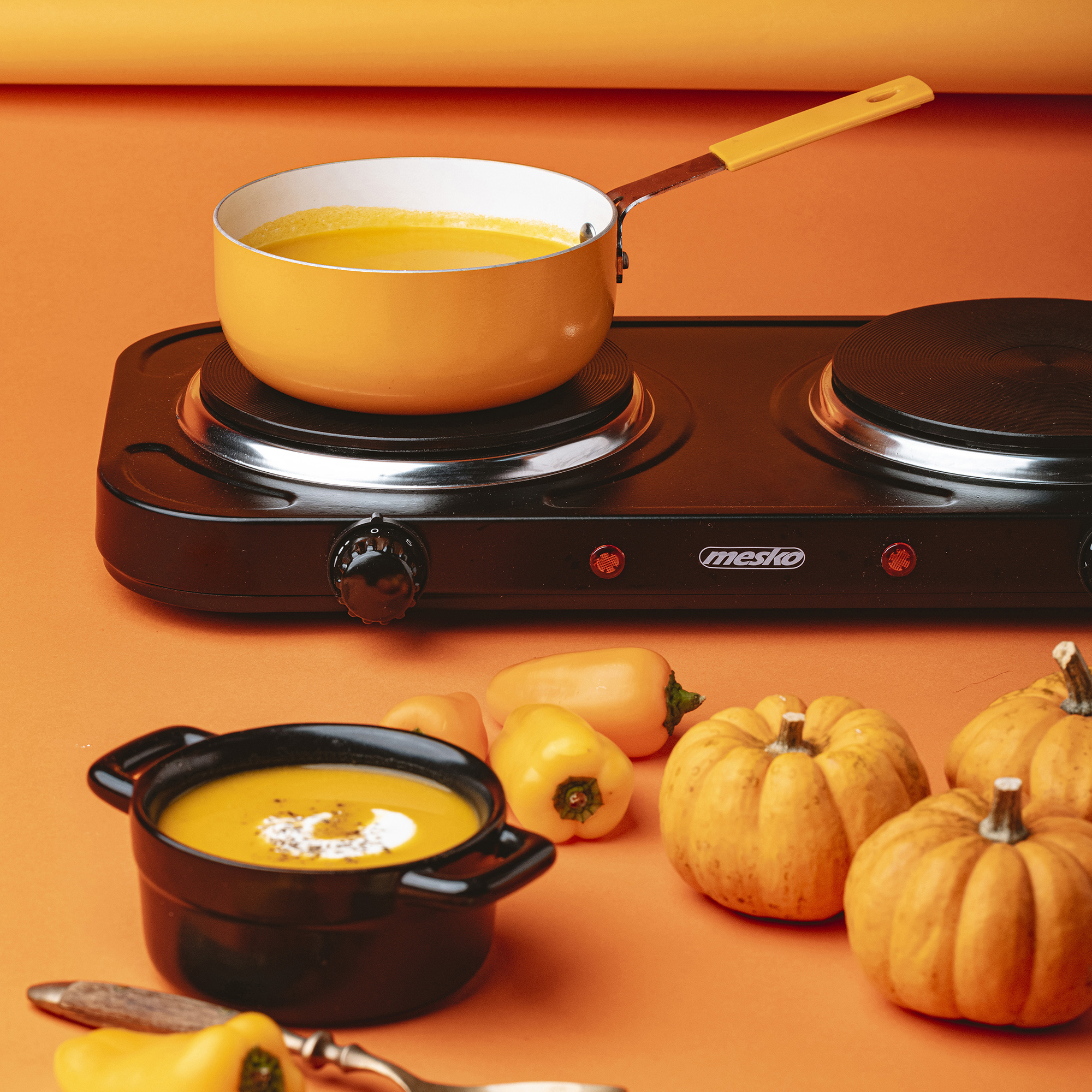 Stół z czarną podwójną płytą grzejną, garnkiem i warzywami na pomarańczowym tle.