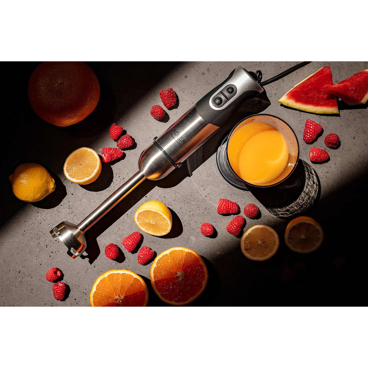 Ein Stabmixer mit Obst: Orange, Zitrone, Himbeeren und ein Glas Saft auf dunkler Oberfläche.
