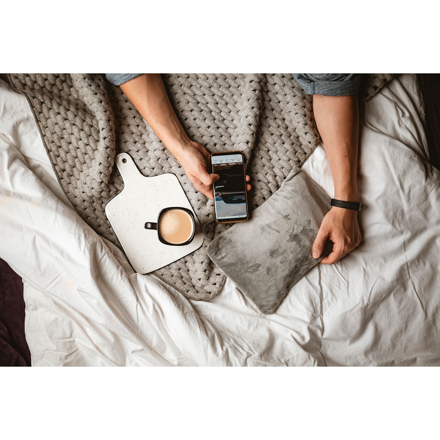 Persona en la cama con teléfono, café y un rastreador de actividad en la muñeca.