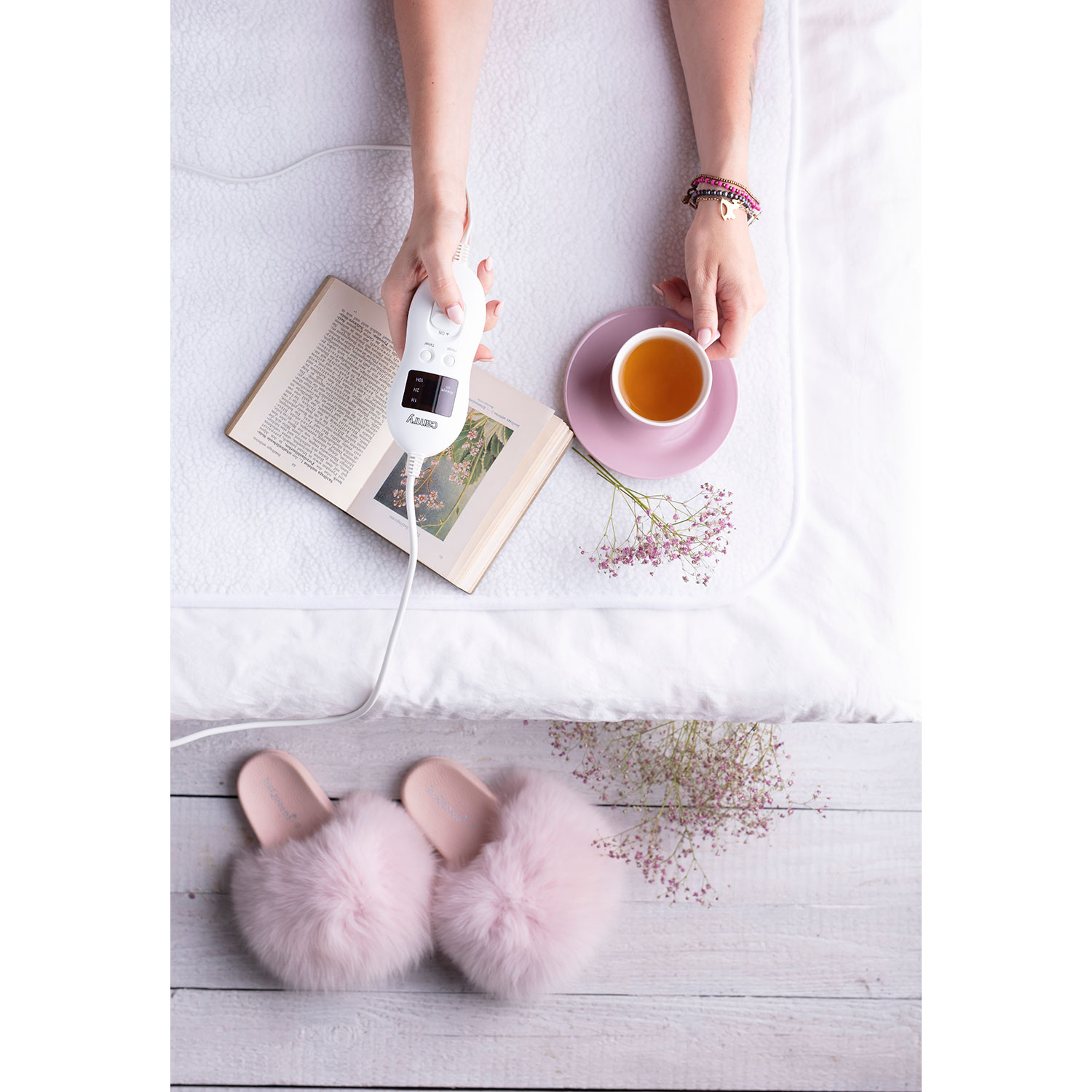 Hände halten ein Bedienfeld über einem Buch und einer Tasse Tee. Rosa Hausschuhe sind in der Nähe.