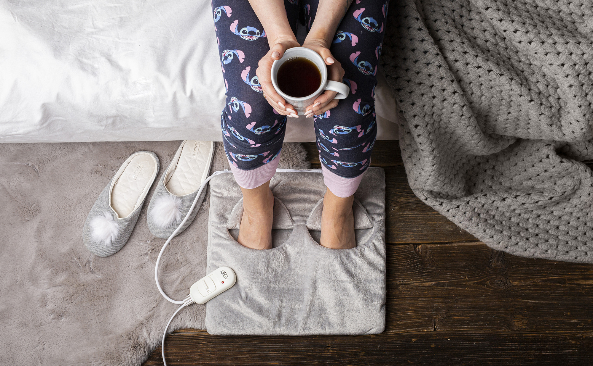 Persona sentada en cama con taza, pies en calientapiés y zapatillas cerca.