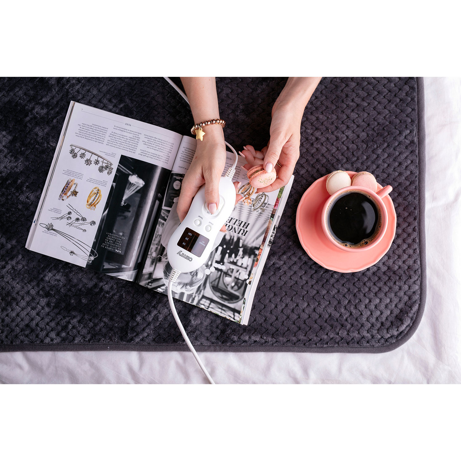 Mani che tengono una rivista e un telecomando. C'è una tazza di caffè e macarons.