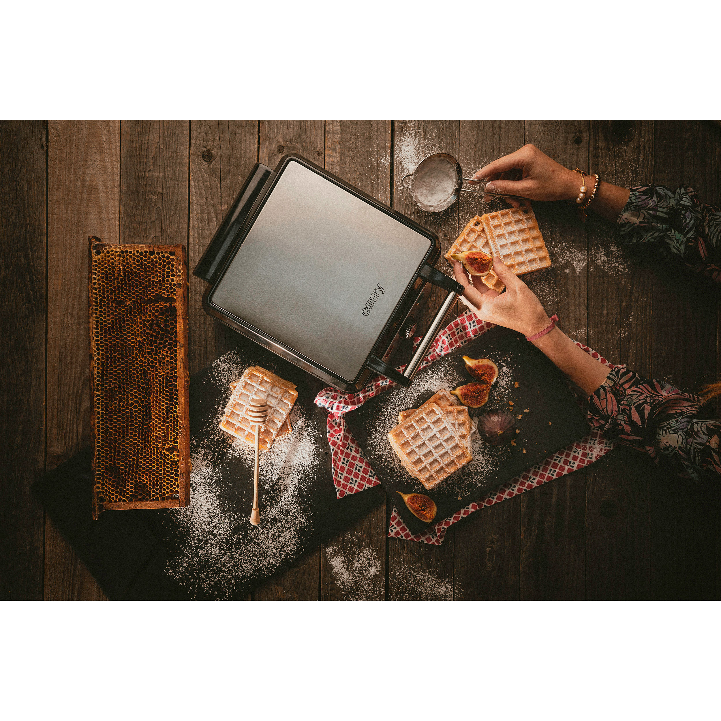 Gofry, miód, figi na drewnianym stole. Dłonie dodają cukier puder. Ciemna drewniana powierzchnia.