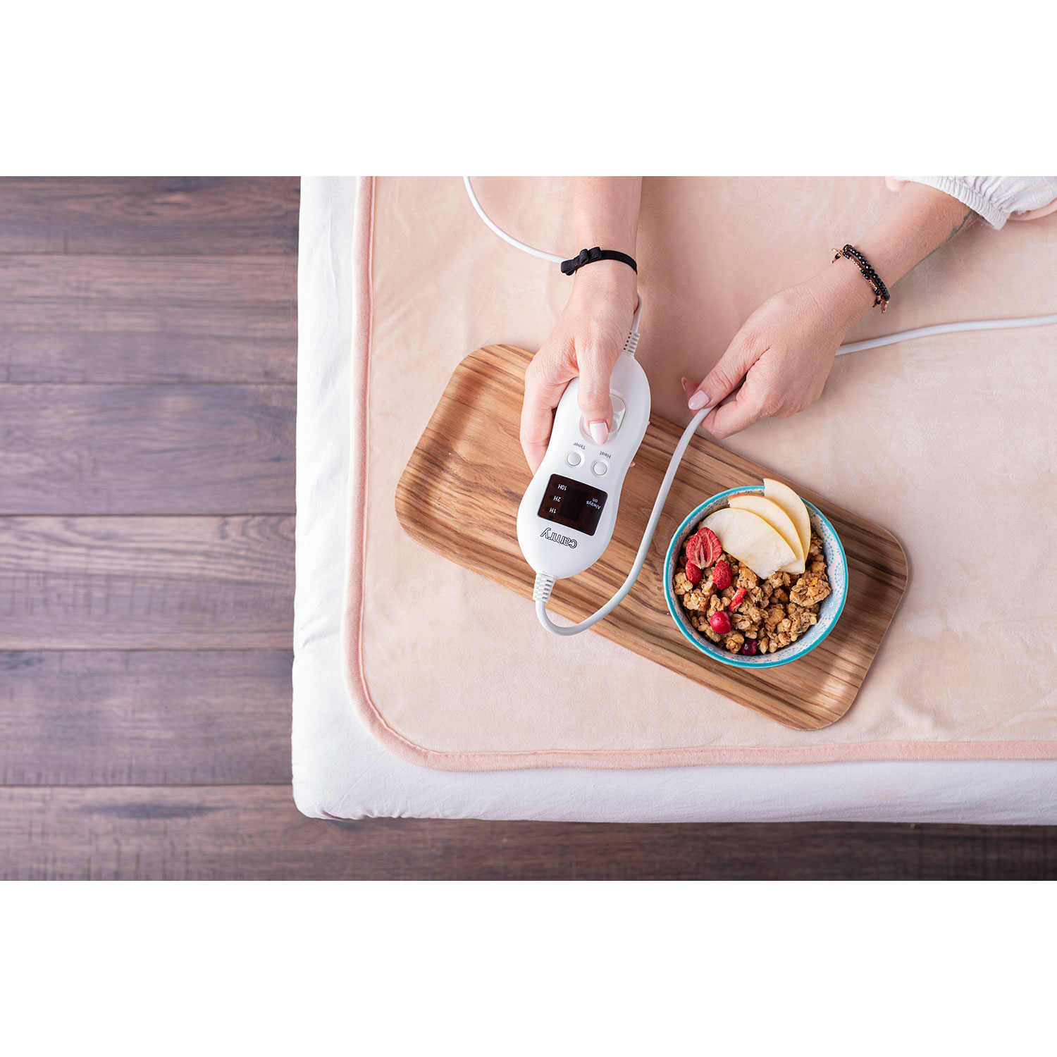 Mujer usando un controlador, bandeja de madera con comida, en una cama. Fondo blanco.