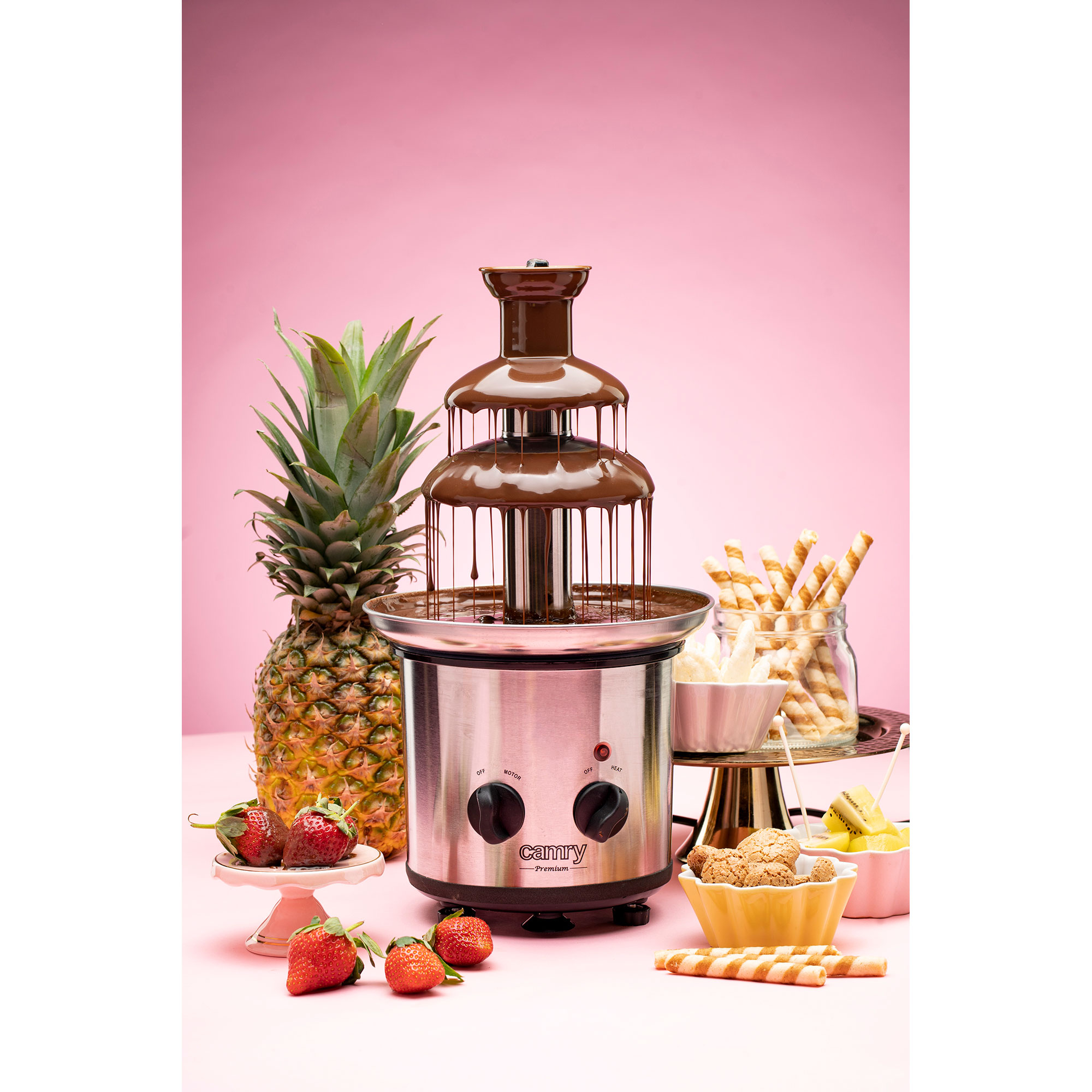 Fuente de chocolate con chocolate fluyendo. Frutas y galletas alrededor, fondo rosa.