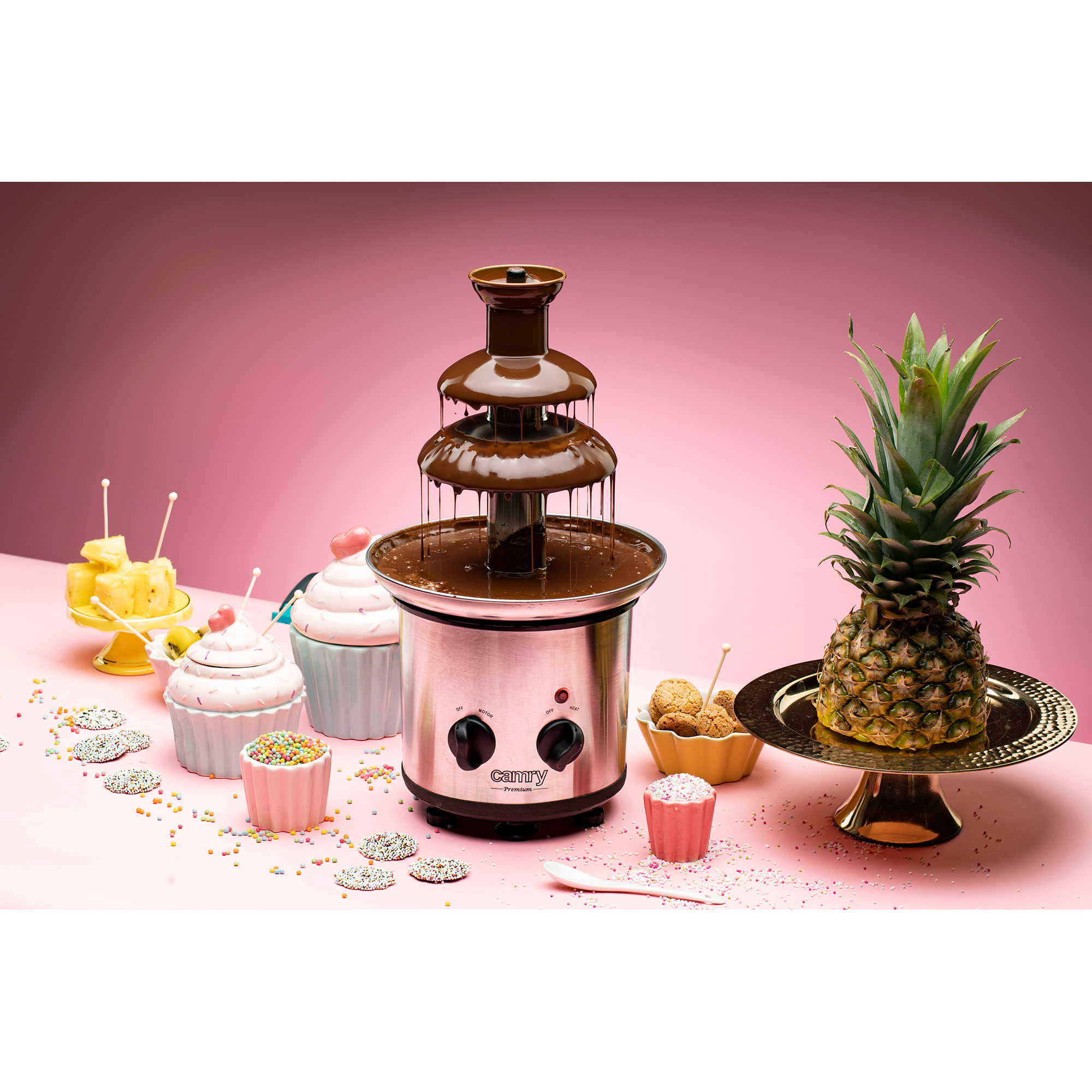 Fuente de chocolate con caramelos, cupcakes y piña sobre fondo rosa.