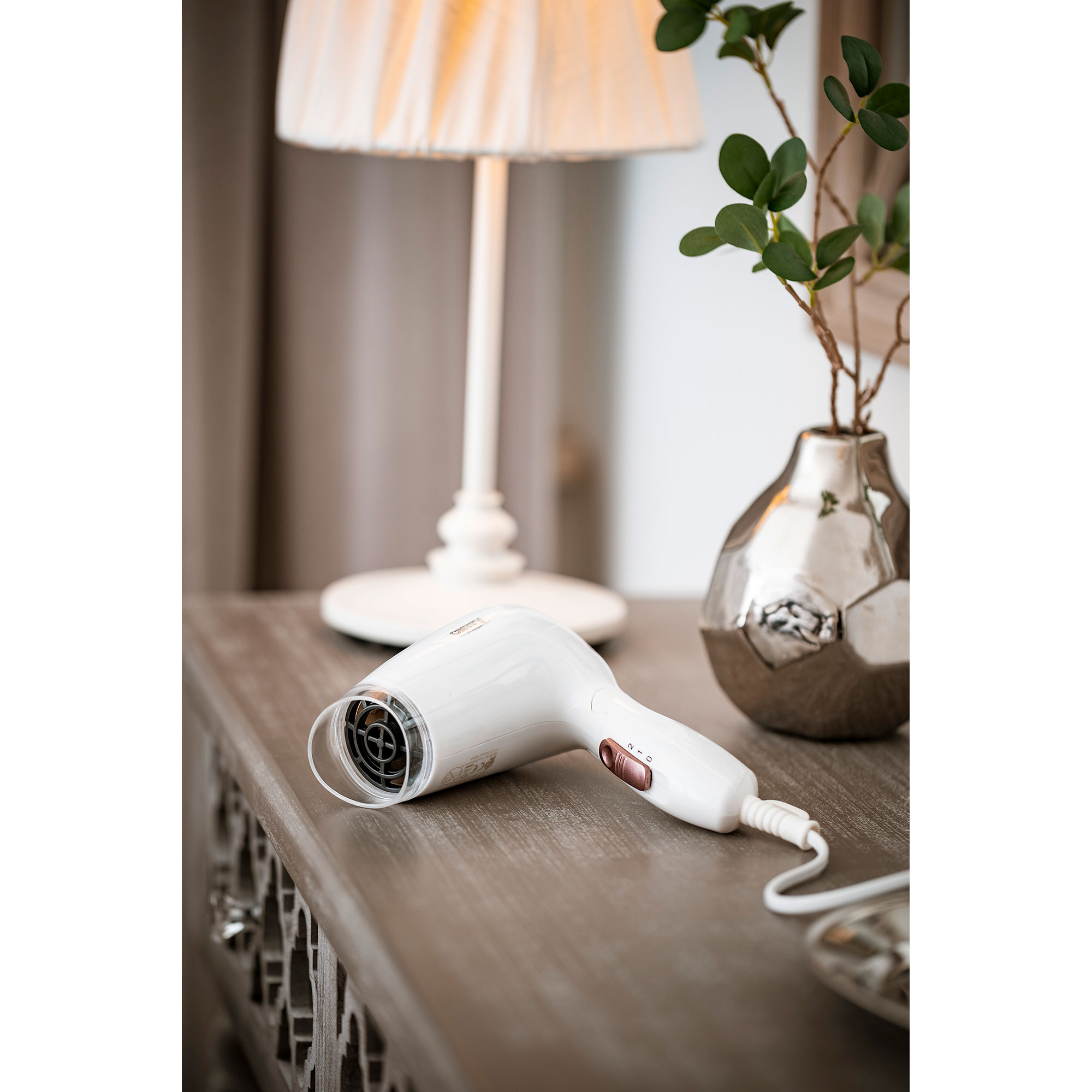 Biała suszarka do włosów na drewnianym stole, z lampą i wazonem w tle.