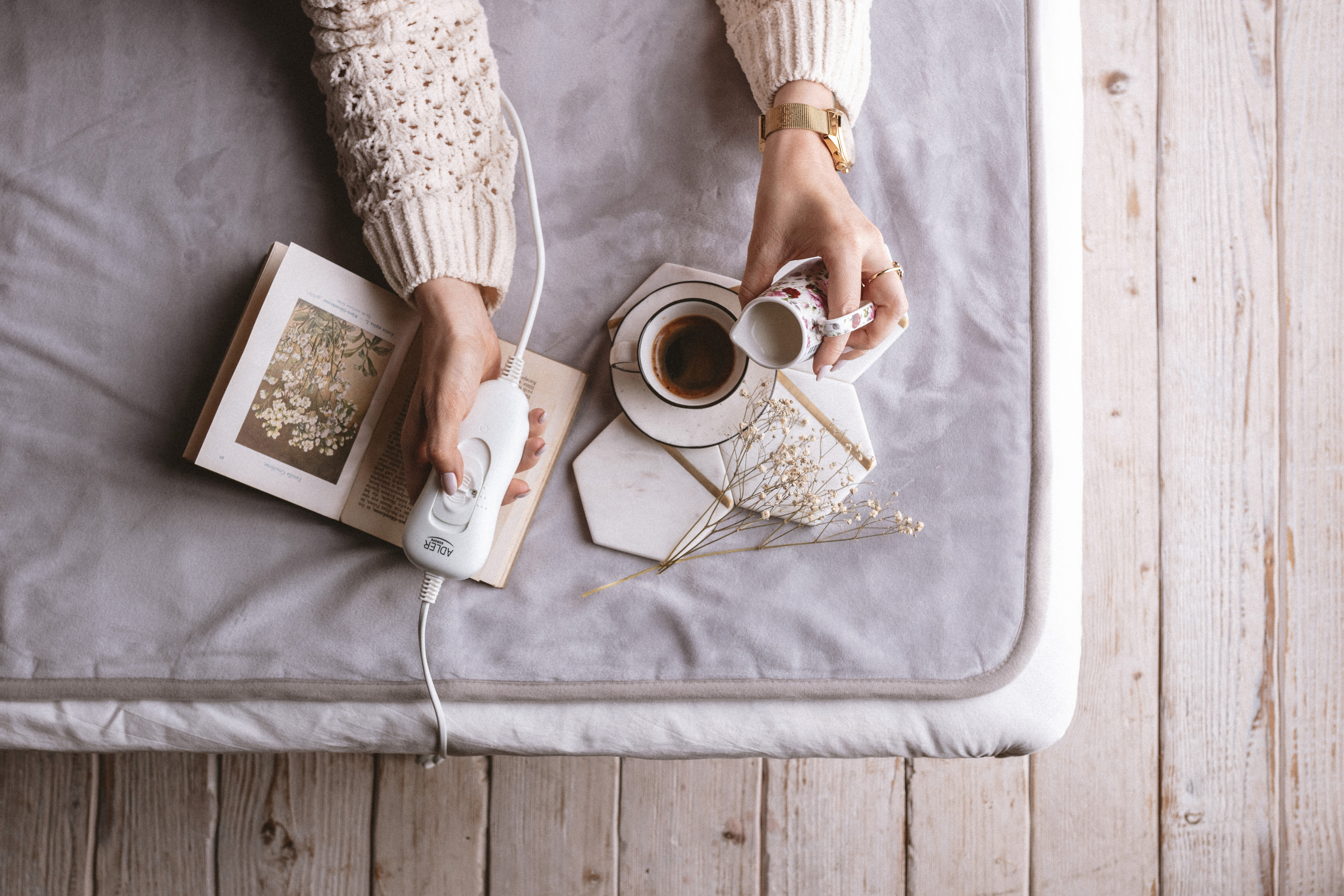 Manos ajustando el control de una almohadilla térmica, con café, libro y flores cerca. Todo sobre una cama.