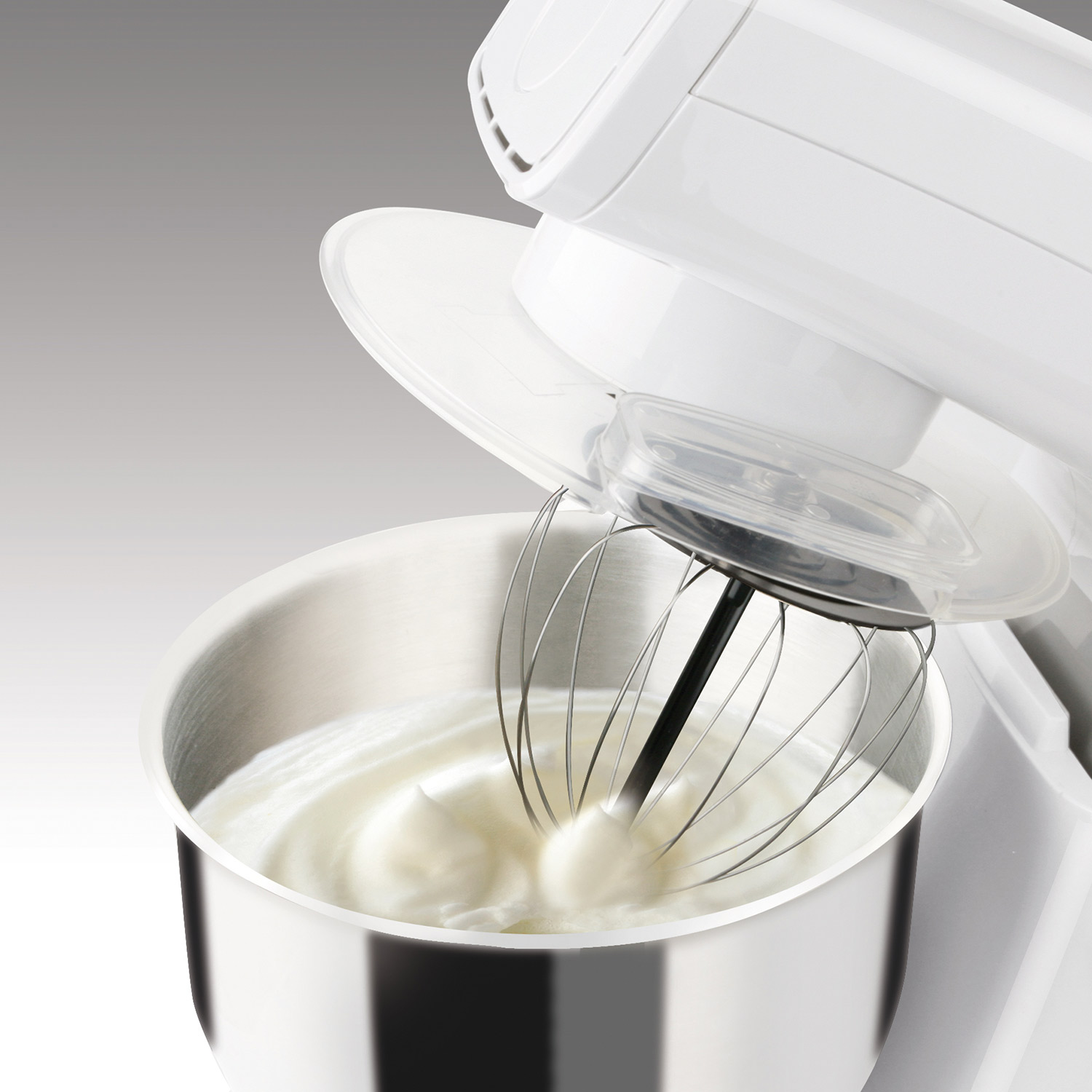 Una batidora blanca bate crema batida en un cuenco de acero inoxidable. La tapa está abierta.