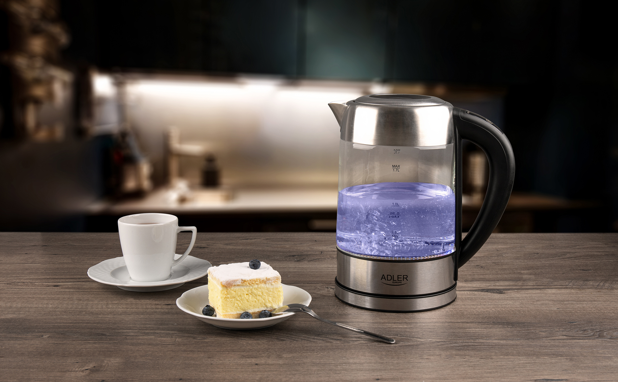 Czajnik elektryczny z wrzącą wodą, filiżanka kawy, kawałek ciasta na talerzu. Drewniany stół i rozmyte tło kuchni.