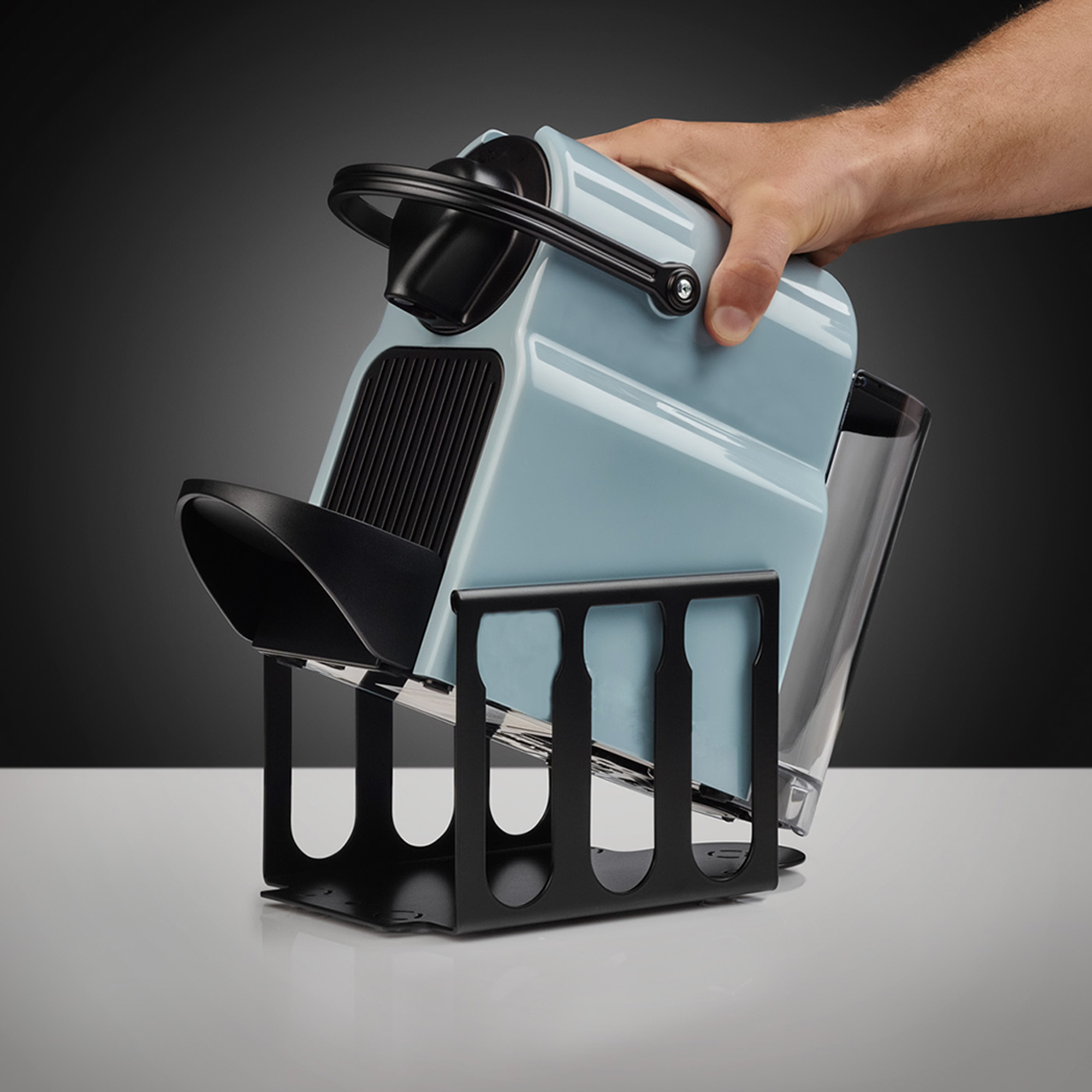 Eine Hand hält eine blau-schwarze Kaffeemaschine über einem schwarzen Ständer. Es gibt einen transparenten Behälter.