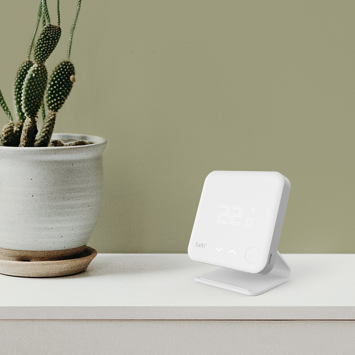 A white smart thermostat with a digital display, placed on a white surface next to a cactus.