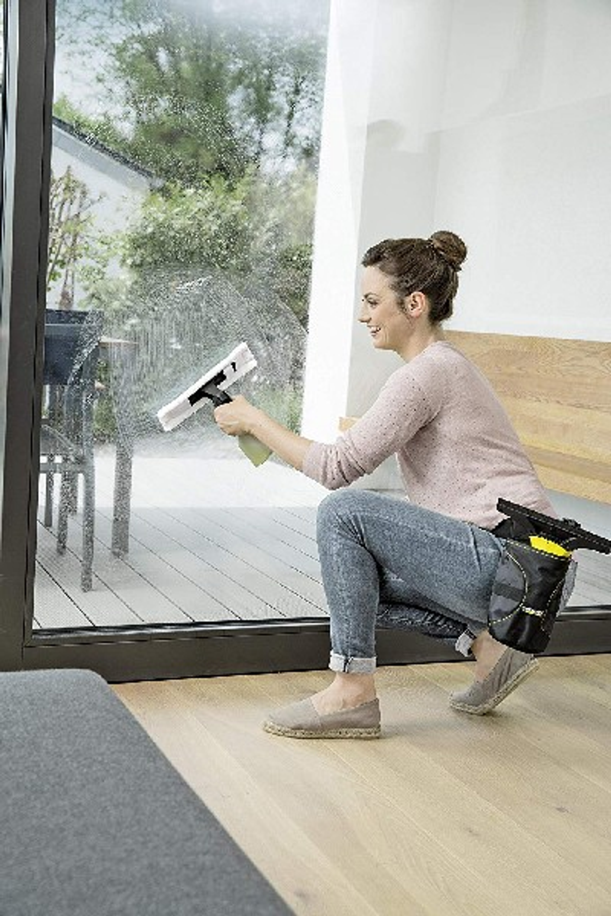 Mujer limpiando una ventana con un pulverizador y una rasqueta. Luz solar y casa al fondo.