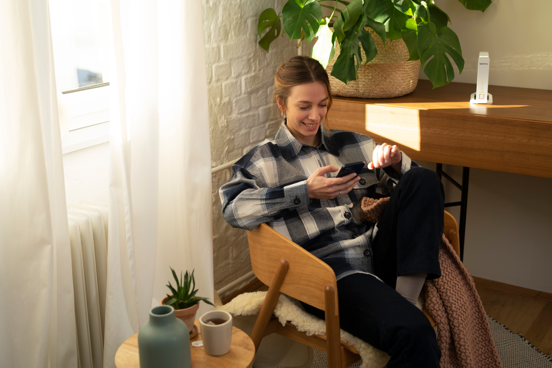 Kobieta siedzi, używa telefonu. Rośliny, okno i drewniany stół w tle.