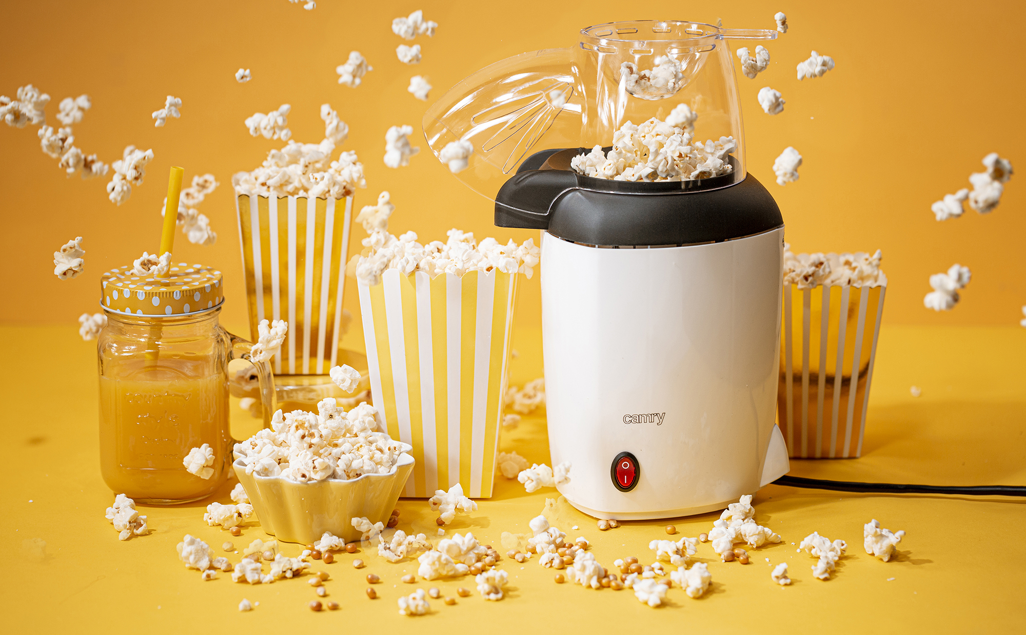 Máquina de palomitas con palomitas reventando y recipientes sobre amarillo.