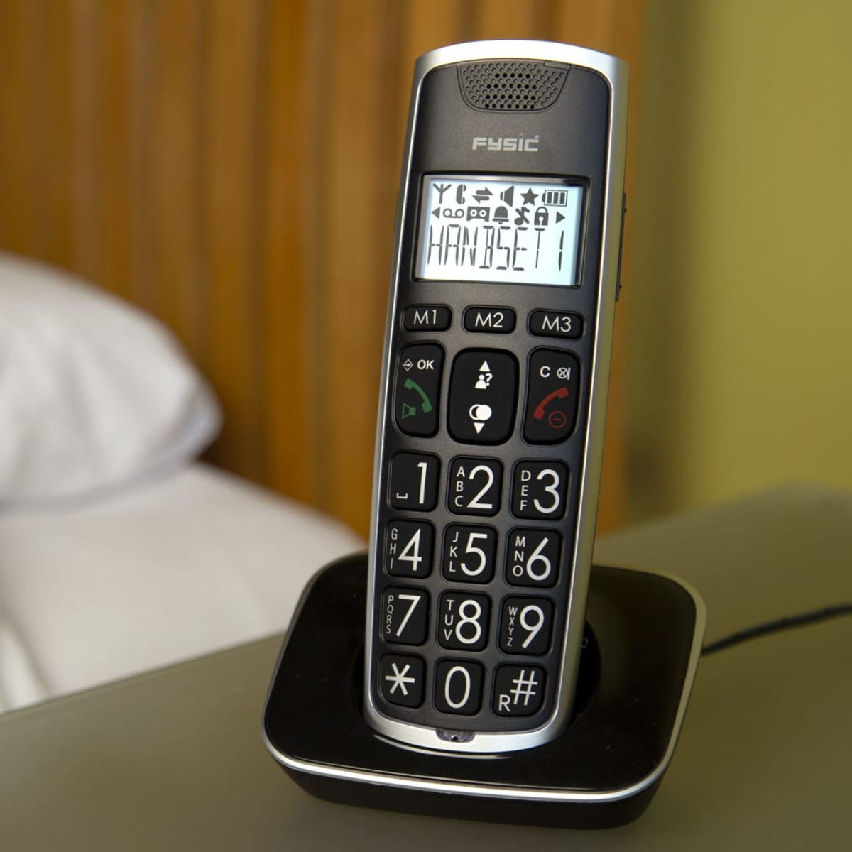 Téléphone sans fil noir et argenté sur socle de chargement, gros boutons. Cadre de chambre.
