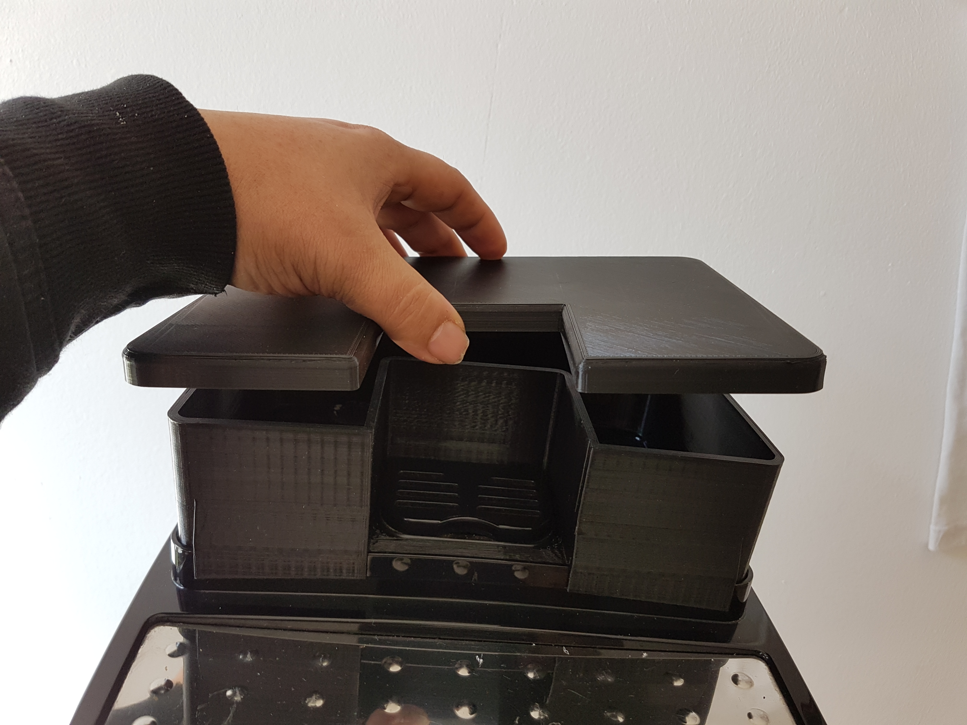 Eine Hand platziert ein schwarzes Plastiktablett in eine größere schwarze Struktur. Die Struktur hat zwei Seitenfächer.