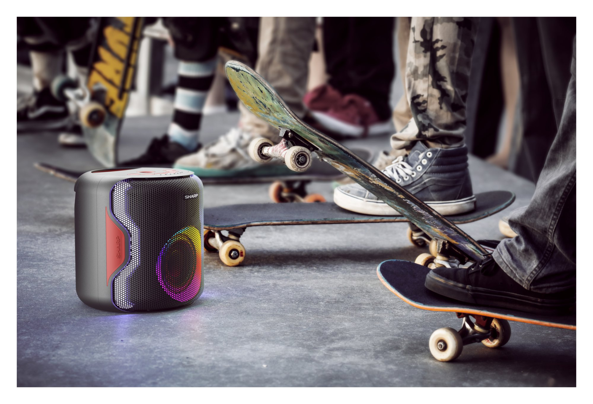 Un altavoz con luces coloridas sobre suelo de hormigón. Personas en monopatines en el fondo.
