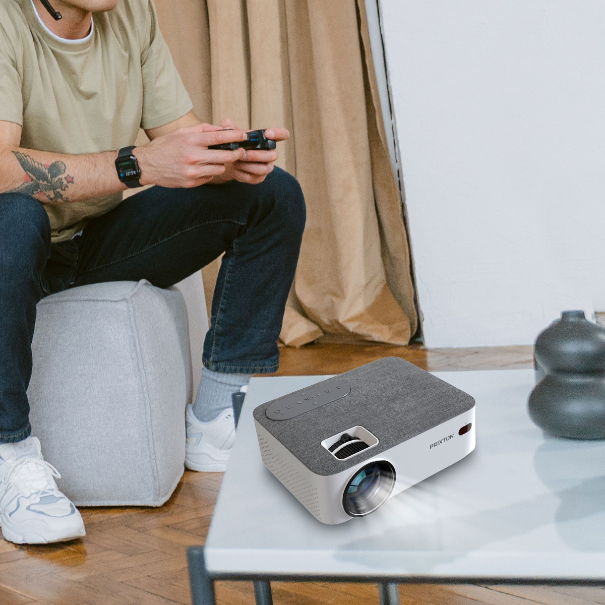 Mężczyzna siedzi na szarej pufie i gra w grę. Projektor stoi na stole.