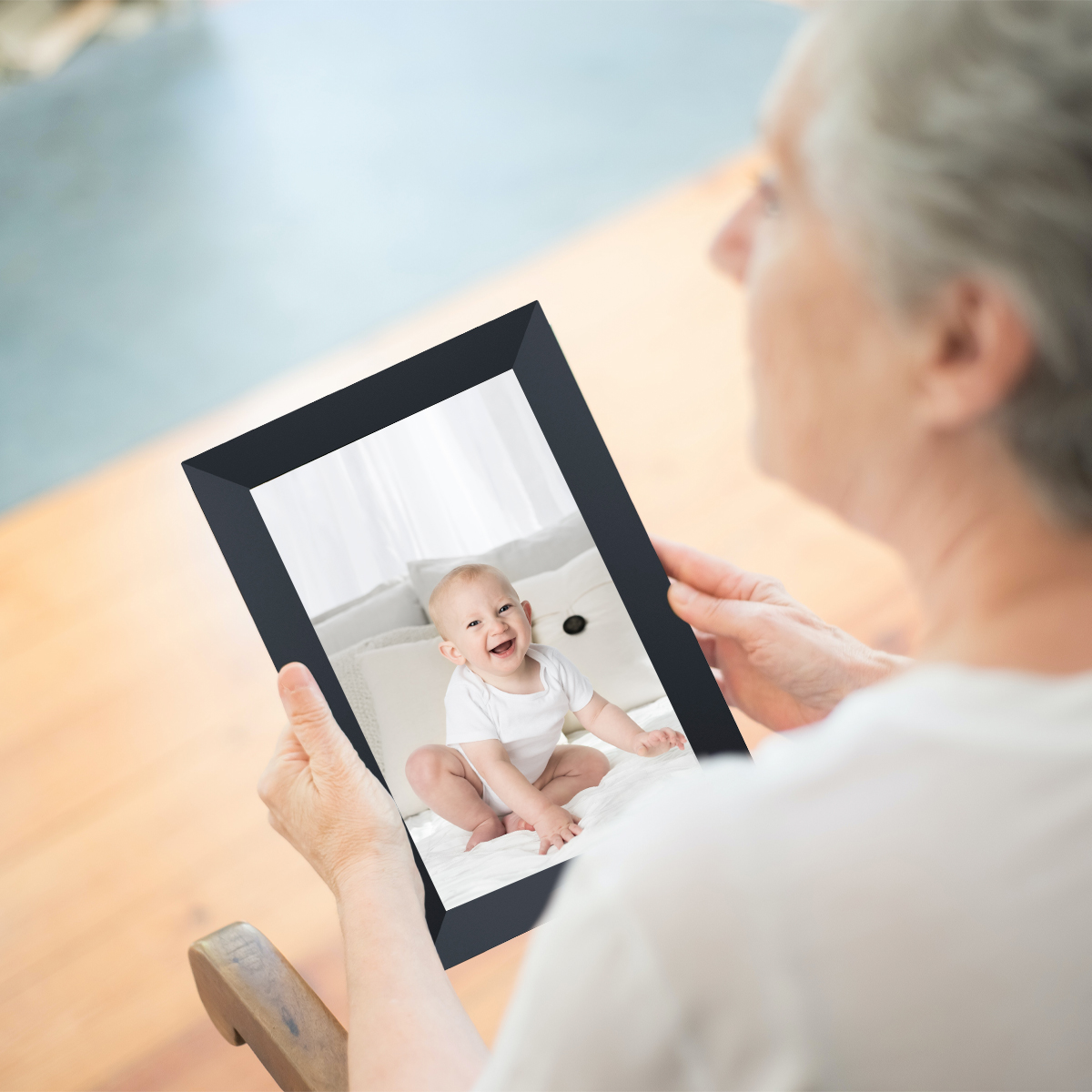 Starsza kobieta trzyma ramkę ze zdjęciem. Zdjęcie uśmiechniętego dziecka. Drewniane krzesło.
