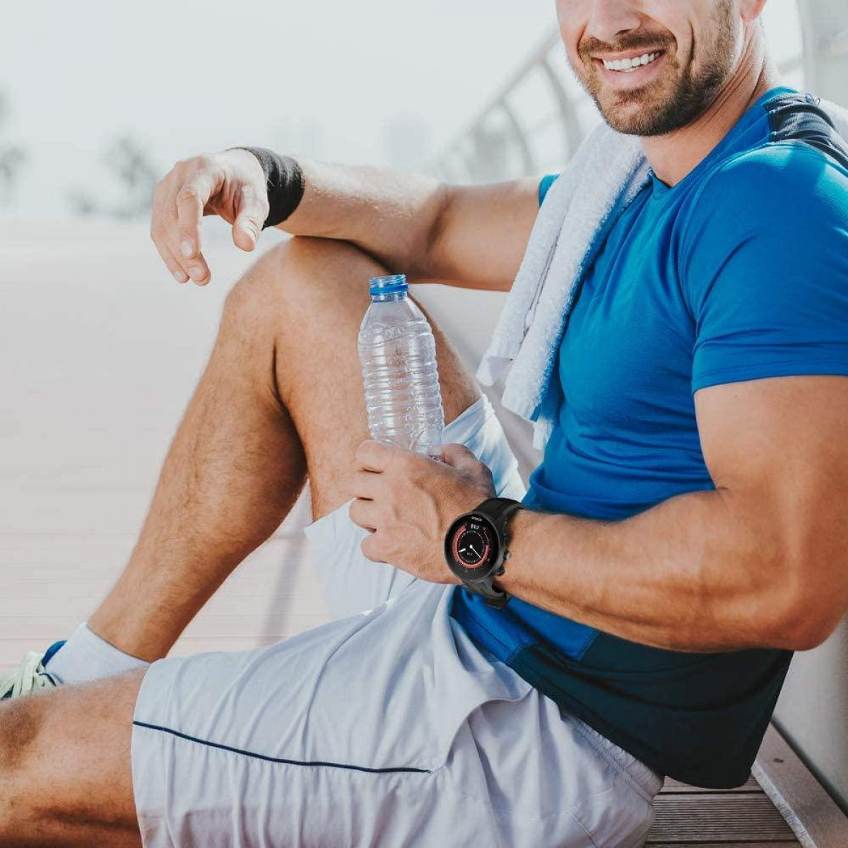 Homme en chemise bleue portant une montre intelligente noire. Il tient une bouteille d'eau et une serviette.