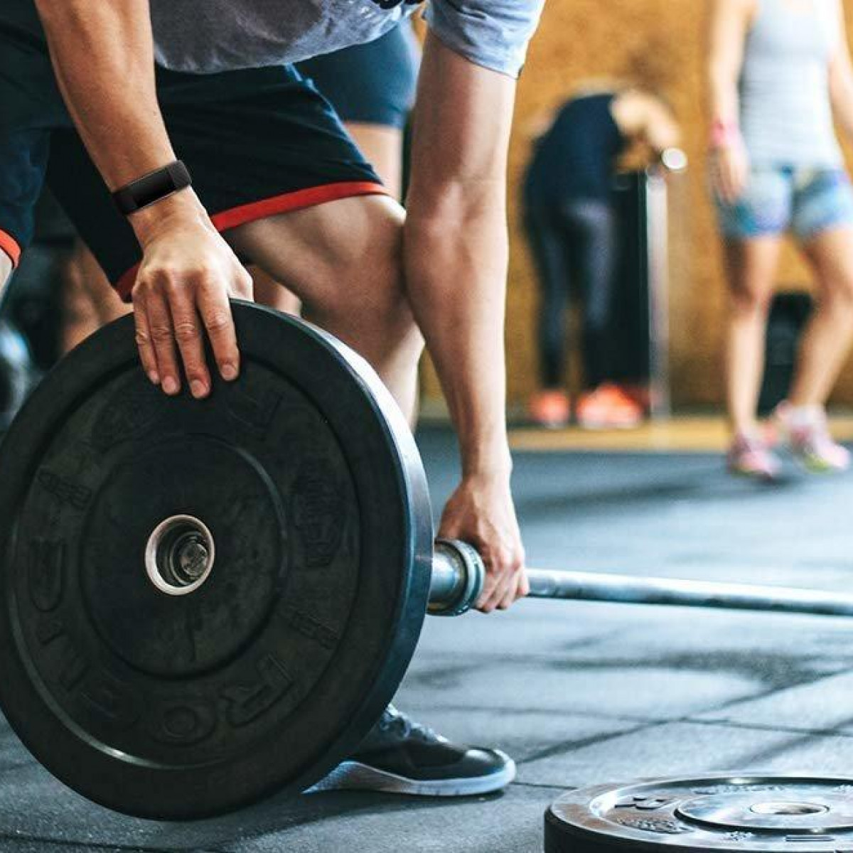 Mann hebt Langhantel mit Gewichten. Gym-Umgebung, andere Personen im Hintergrund unscharf.