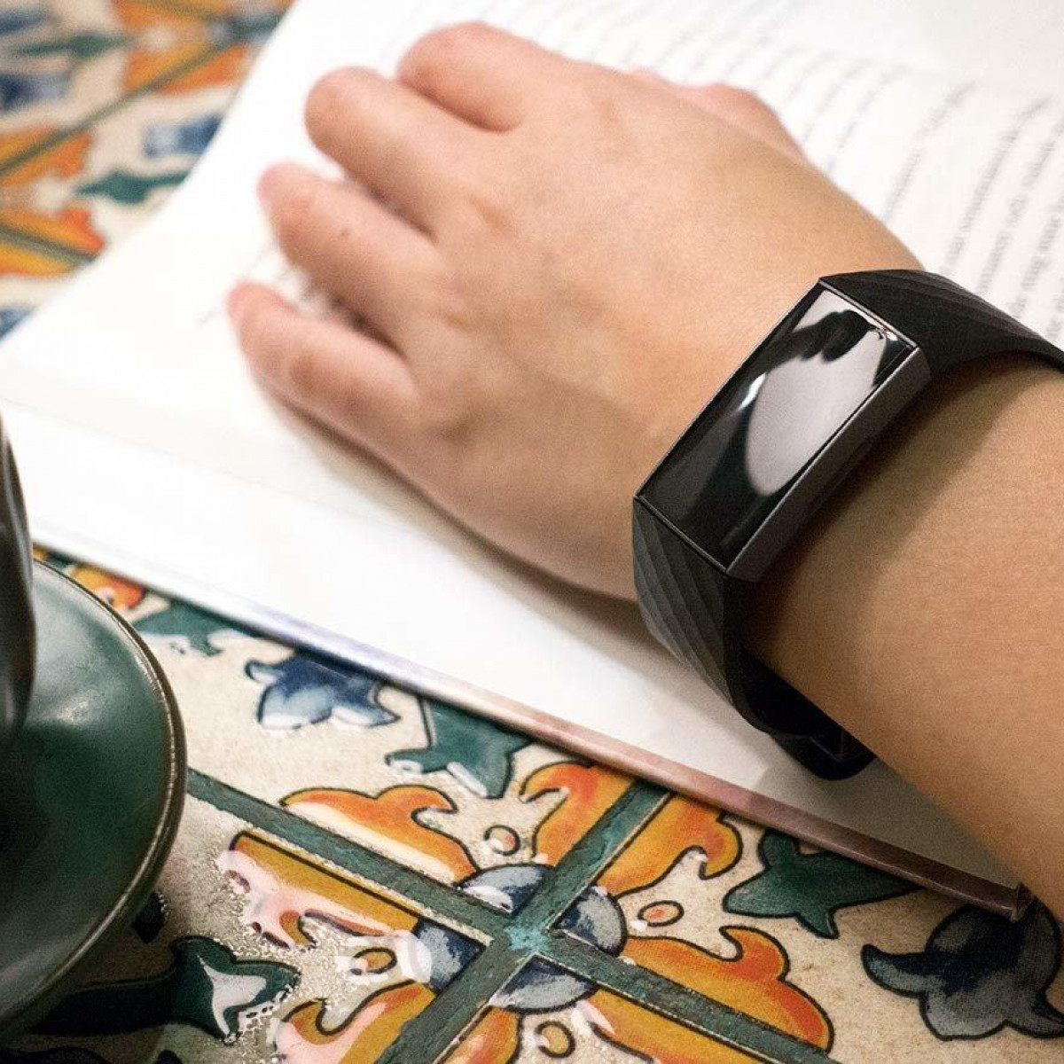 Die Hand einer Person mit einem schwarzen Fitness-Tracker am Handgelenk, die ein Buch liest, mit einer Tasse.