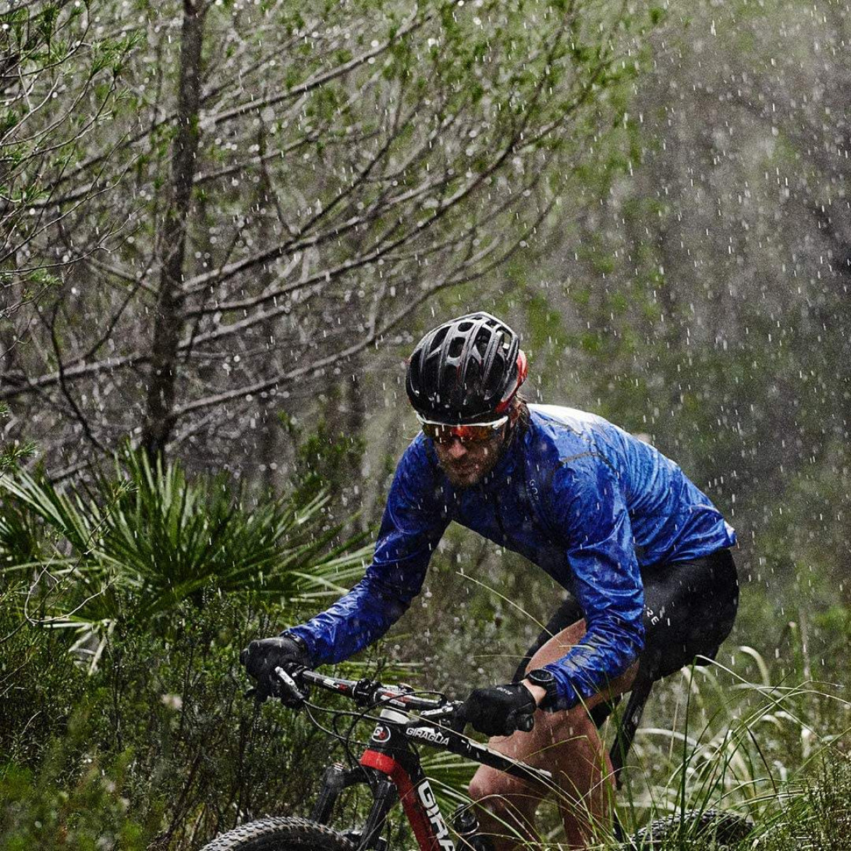 Radfahrer im Regen mit Helm und blauer Jacke, der durch einen Wald fährt.