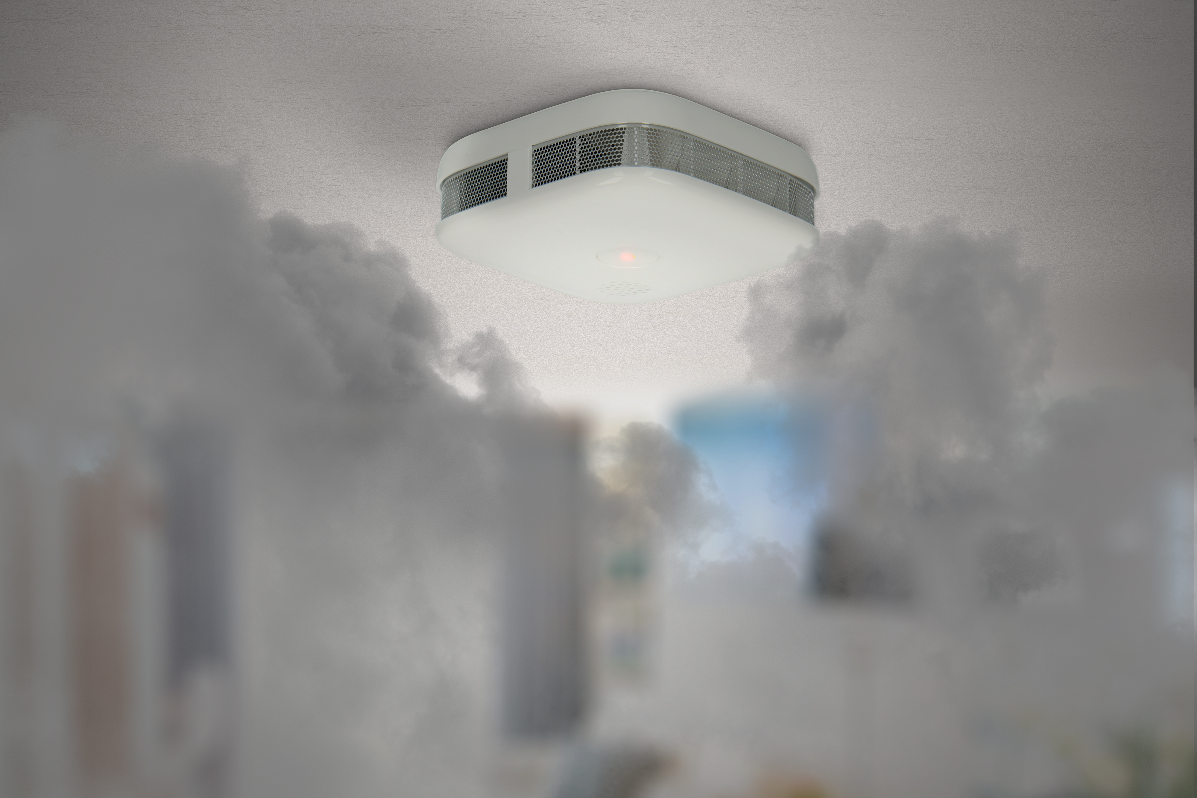Biały czujnik dymu zamontowany na białym suficie, z rozmytym dymem na pierwszym i drugim planie.
