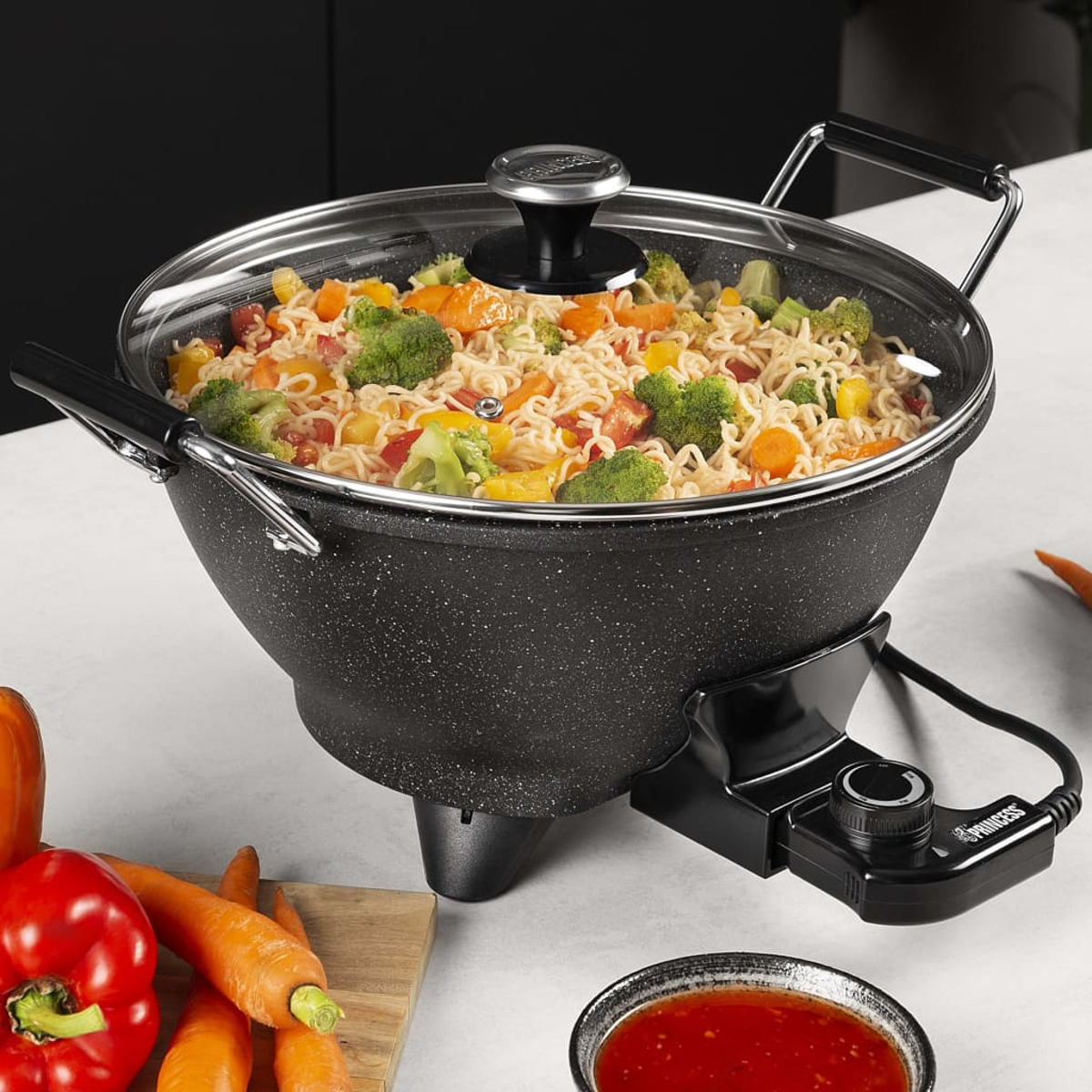 Ein Wok mit Nudeln, Gemüse und Glasdeckel. Der Wok steht auf schwarzer Basis mit Bedienelementen.