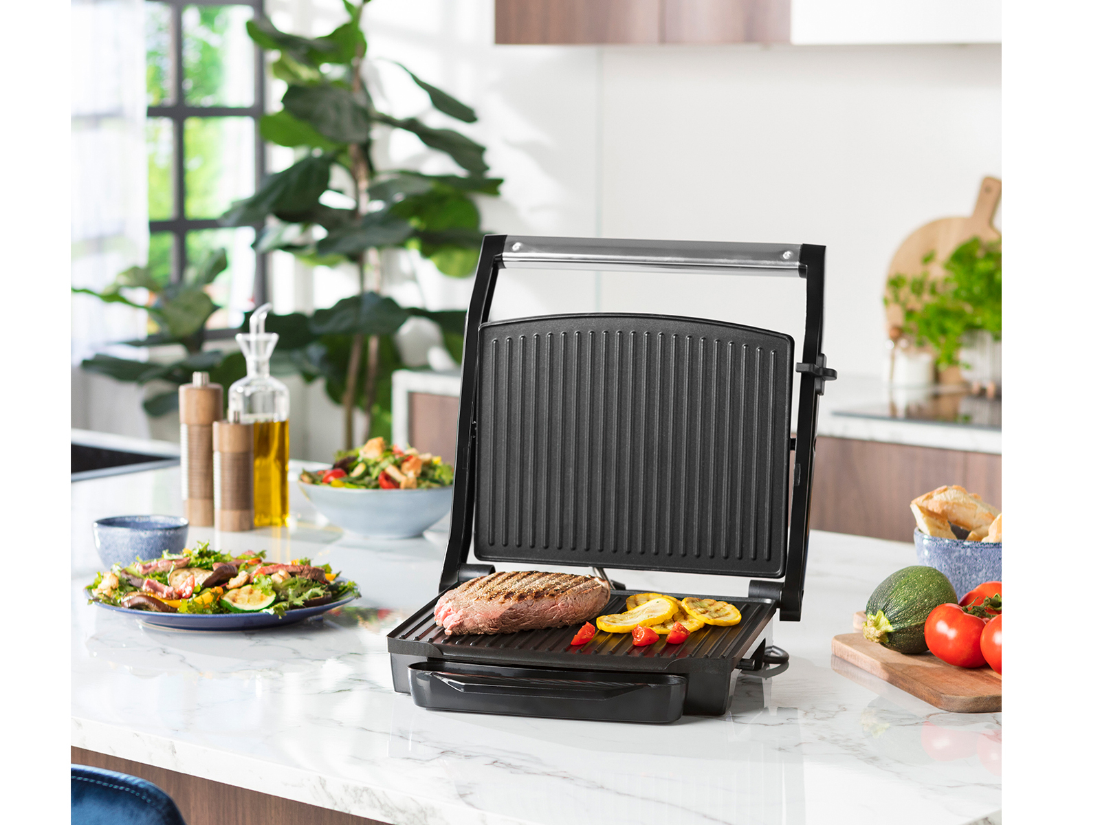 Offener Grill mit Steak, Gemüse und Beilagen auf einer weißen Arbeitsplatte. Verschwommener Hintergrund.