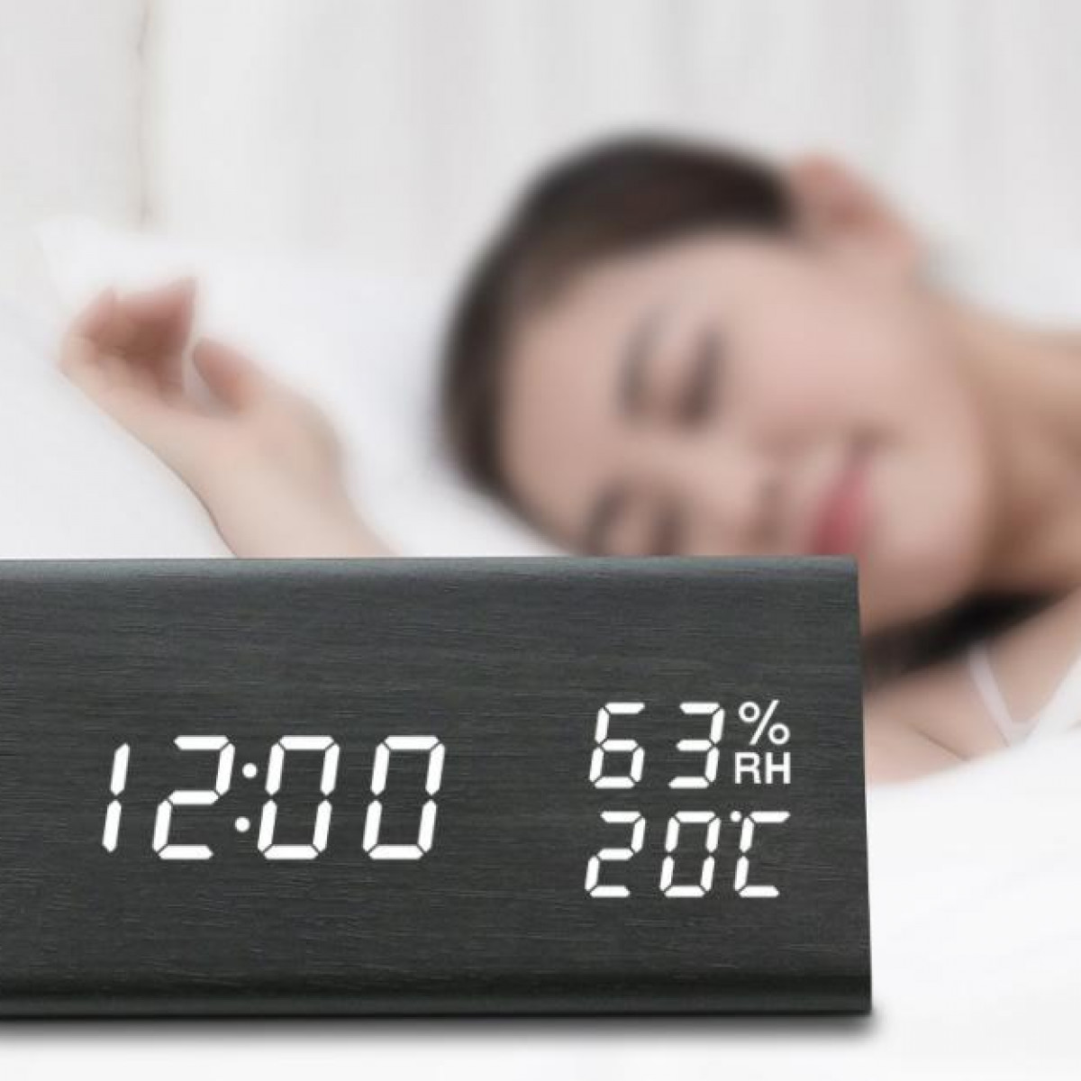 Zwarte digitale houten klok met de tijd 12:00, met een slapende vrouw op de achtergrond.