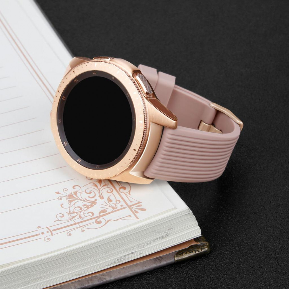 Roségoldene Smartwatch mit schwarzem Bildschirm und rosa Armband auf Notizbuch.